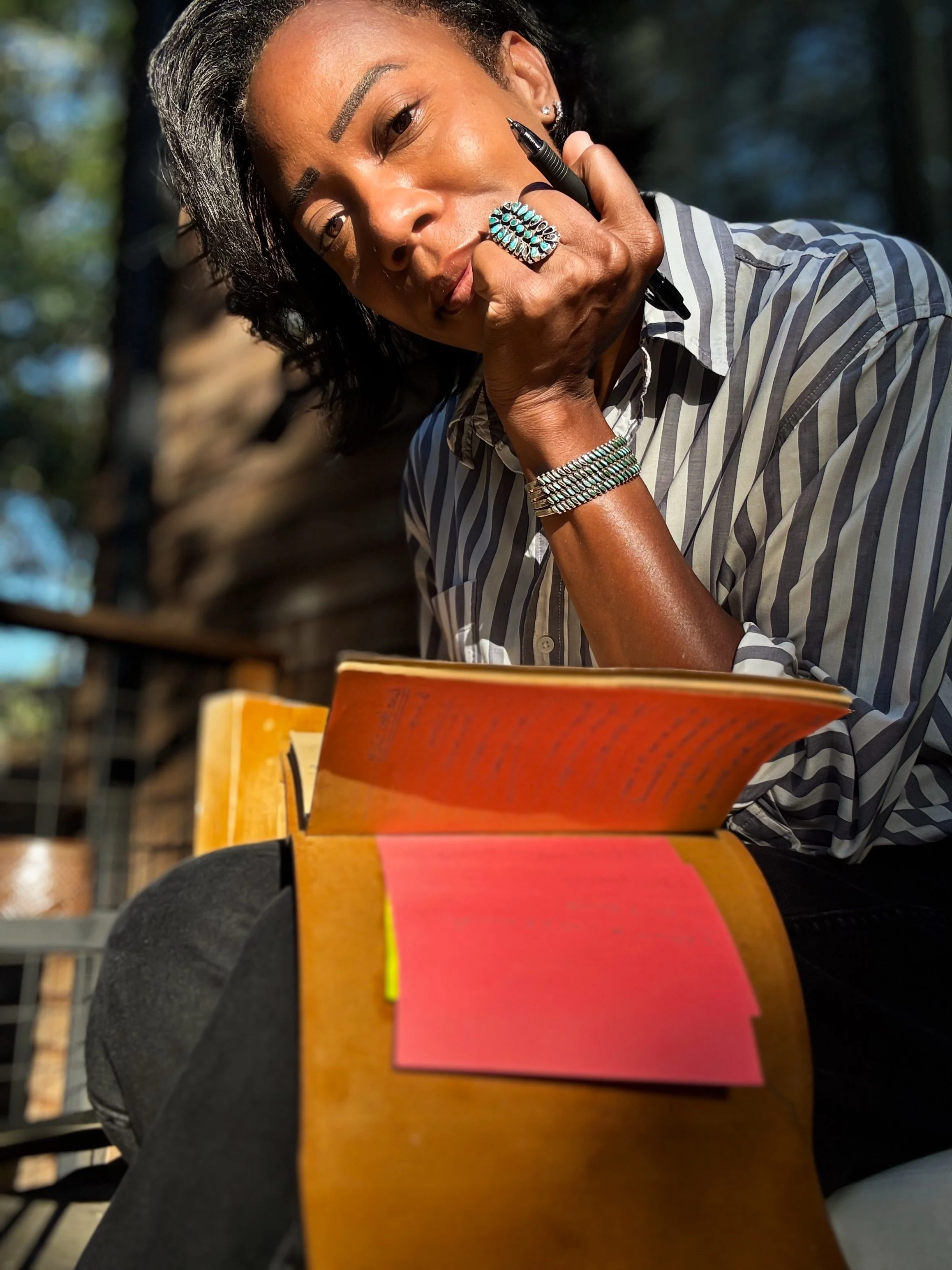 A woman with dark hair, wearing a striped shirt and jewelry, is sitting outdoors reading a book, with sunlight illuminating her face.