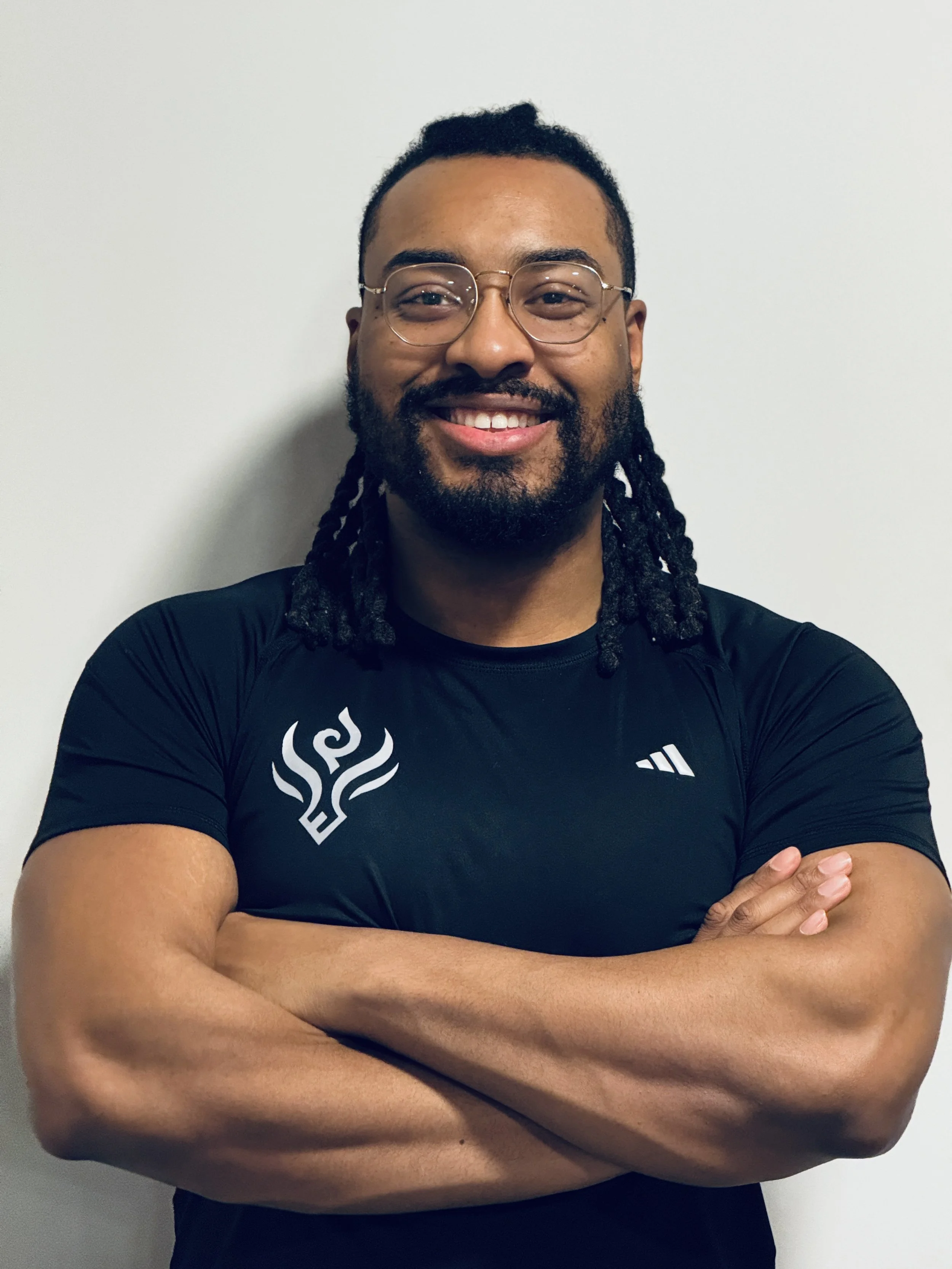 A smiling man with glasses and dreadlocks wearing a black shirt with white designs, crossing his arms in front of a plain light-colored wall.