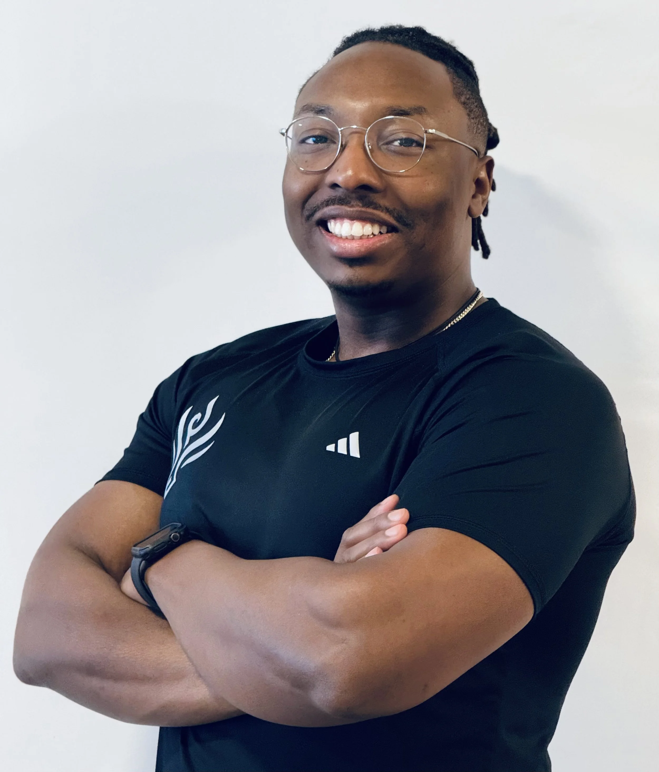 A young man with glasses smiling, wearing a black shirt, a black watch, and a gold chain, standing against a plain white background.