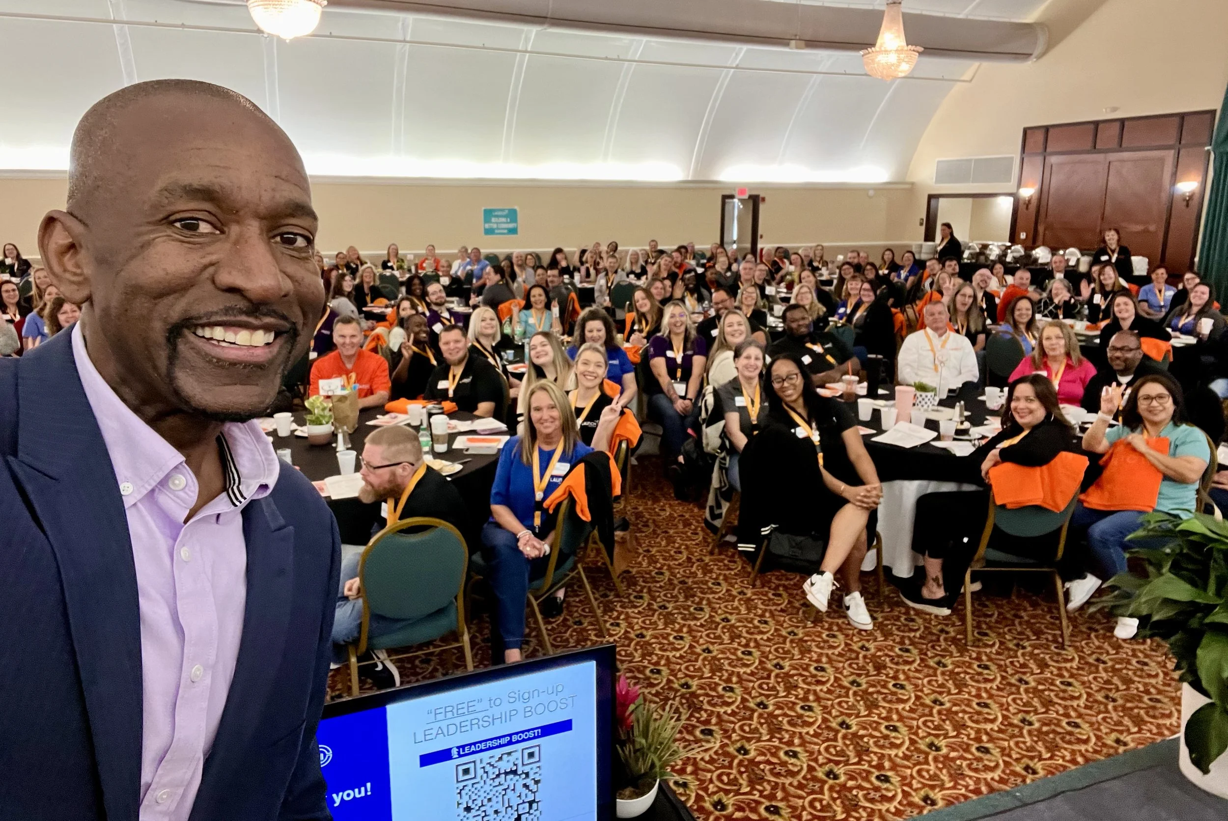 A man taking a selfie at a conference with many attendees seated at tables in the background. The room has a high, curved ceiling, and some attendees are wearing name tags. The man is smiling, dressed in a dark suit and light purple shirt.