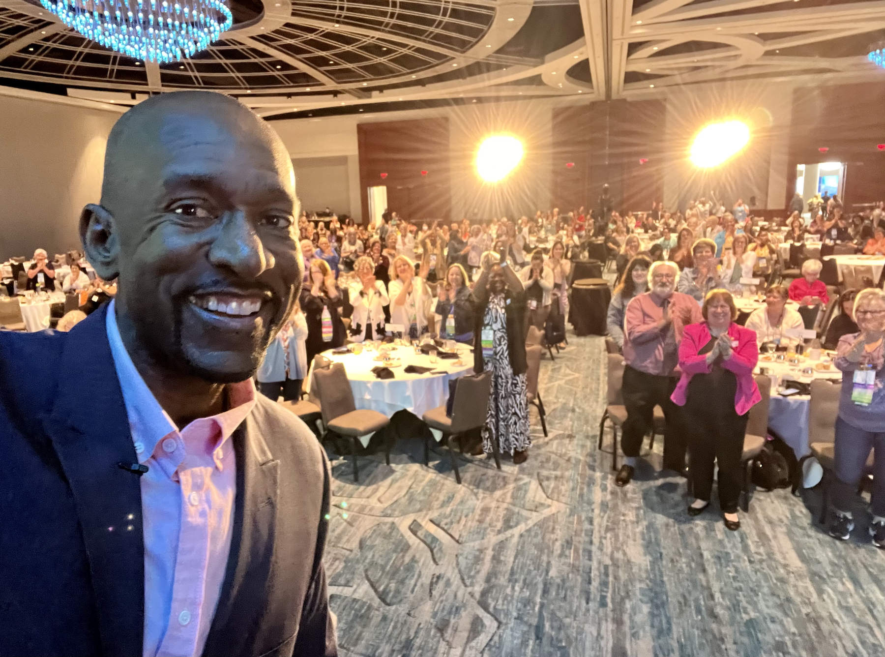 Leadership Speaker Darryl Ross opening keynote for large indoor event with many seated and standing attendees, some clapping, in a banquet hall with round tables and bright lights.