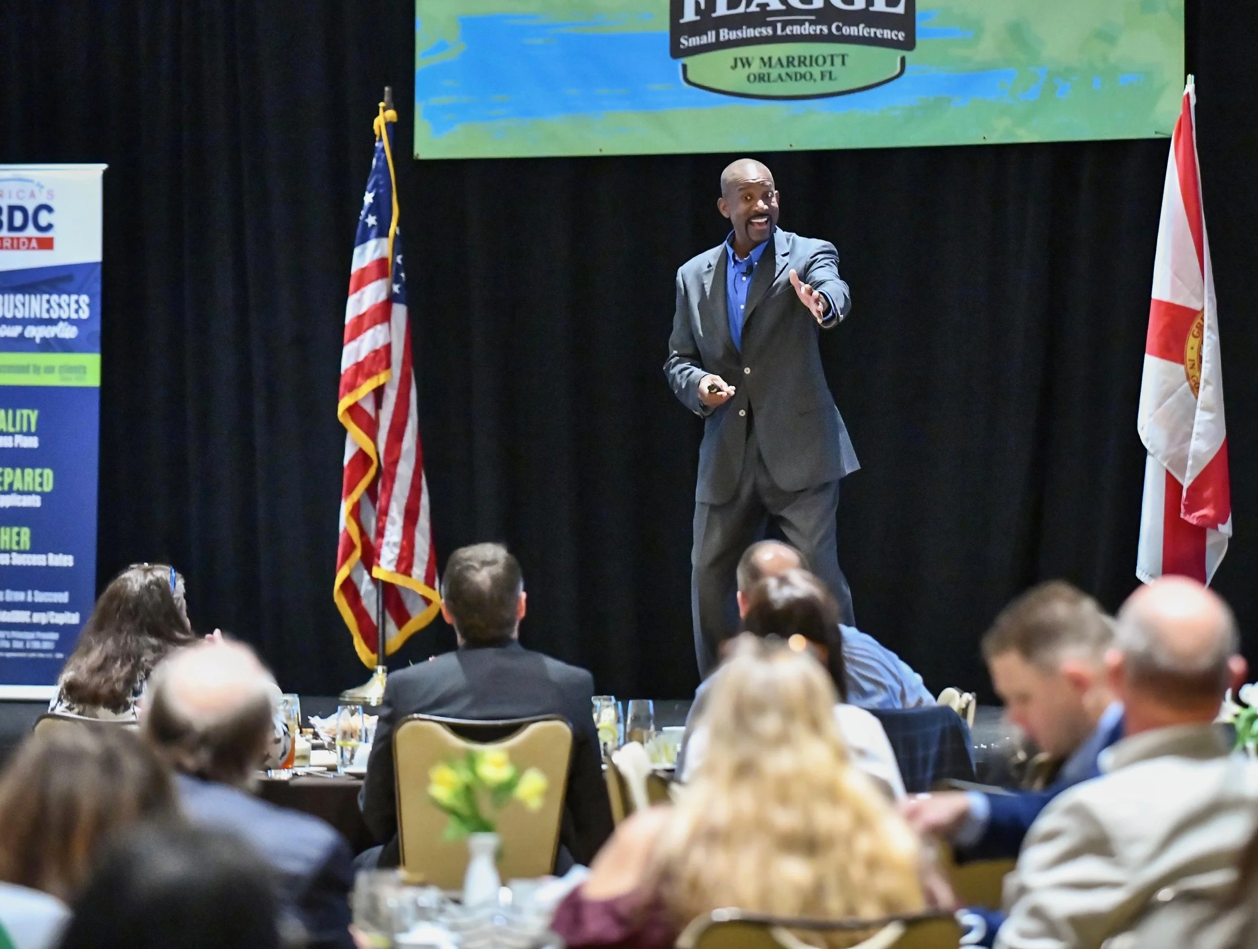 A man in a suit giving a presentation at a conference, with flags on either side and a large screen behind him displaying the conference name. Audience members are seated at tables in the foreground.