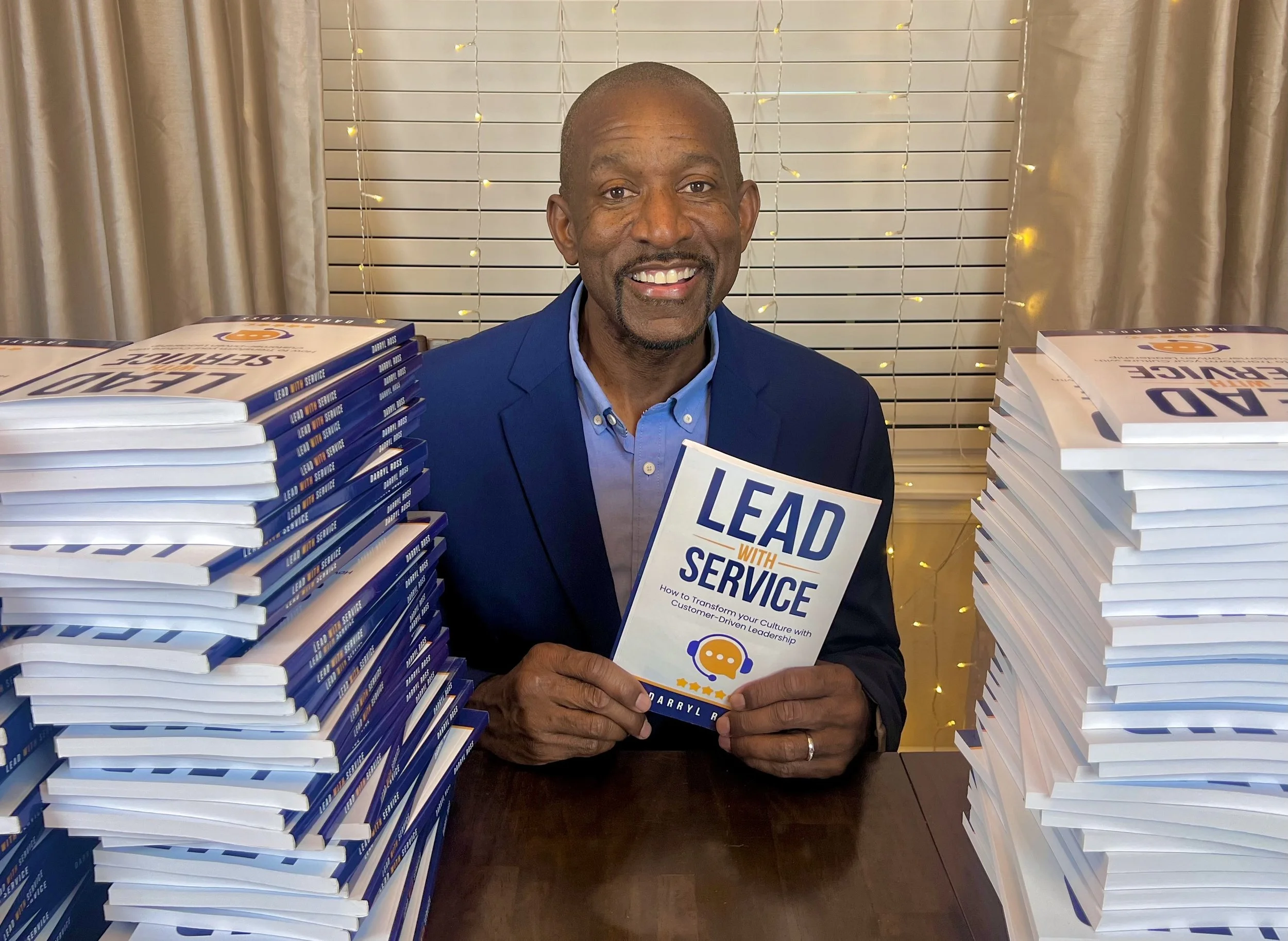 A smiling man in a blue suit holding a book titled 'Lead with Service' surrounded by stacks of the same book, with string lights and blinds in the background.