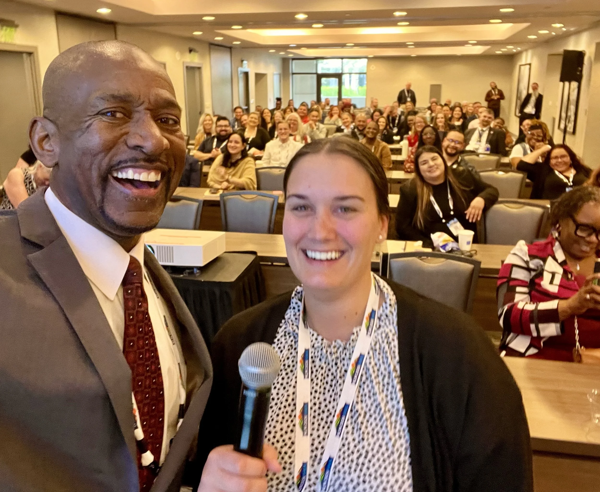 A group of conference attendees in a room, with two people at the front taking a selfie. The man on the left is smiling and wearing a suit and tie, and the woman on the right, holding a microphone, is also smiling. The background shows a large room filled with seated people, many smiling and looking at the camera.