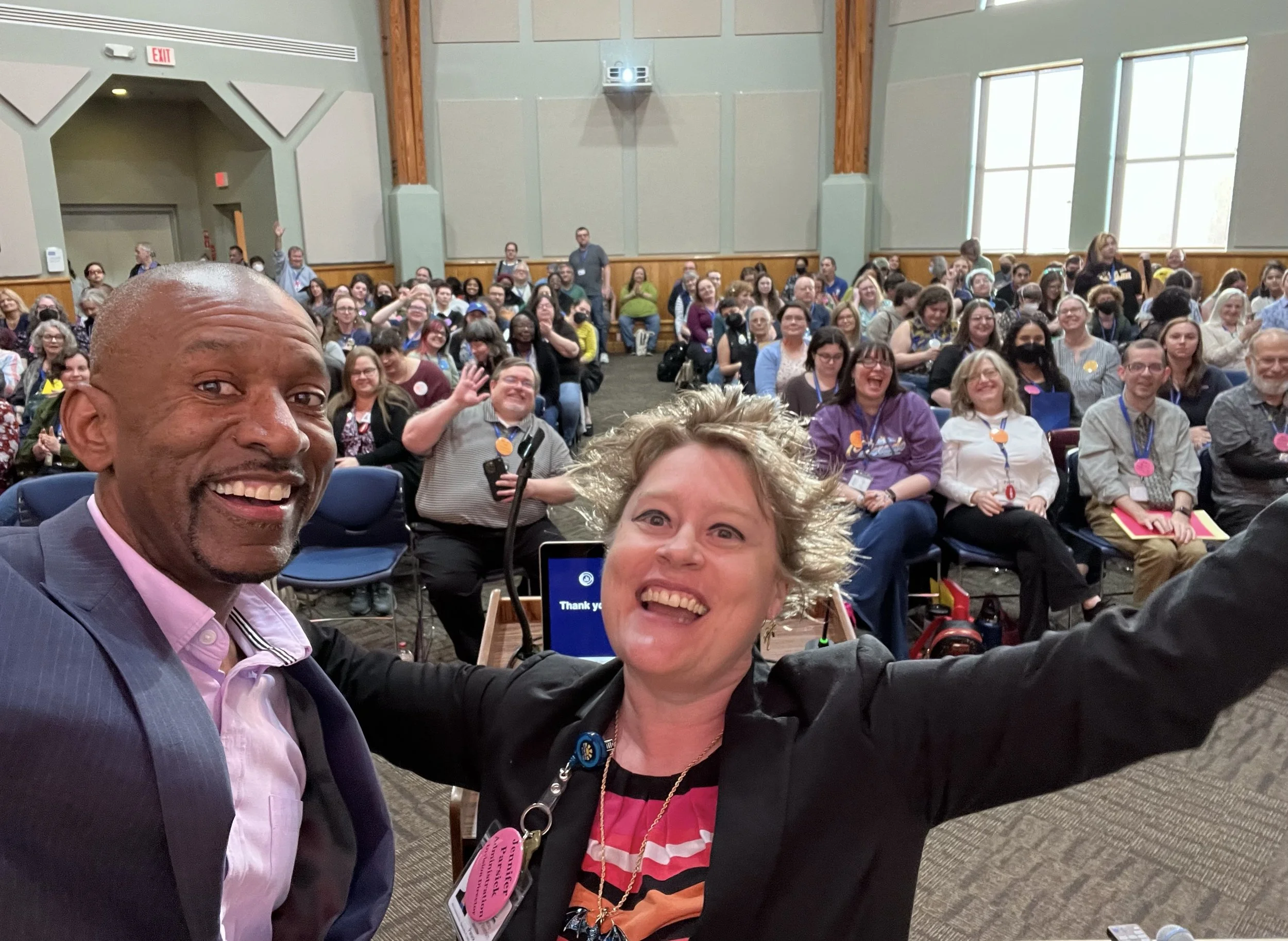 A group of people attending a conference or seminar, smiling and looking at the camera for a selfie, with some waving. The image includes men and women of diverse ages, seated in a large room with high windows and acoustic panels on the walls. The foreground features three individuals, with a man in a suit and two women, one with curly blonde hair and the other with glasses. The room is filled with people, many wearing conference badges or lanyards.