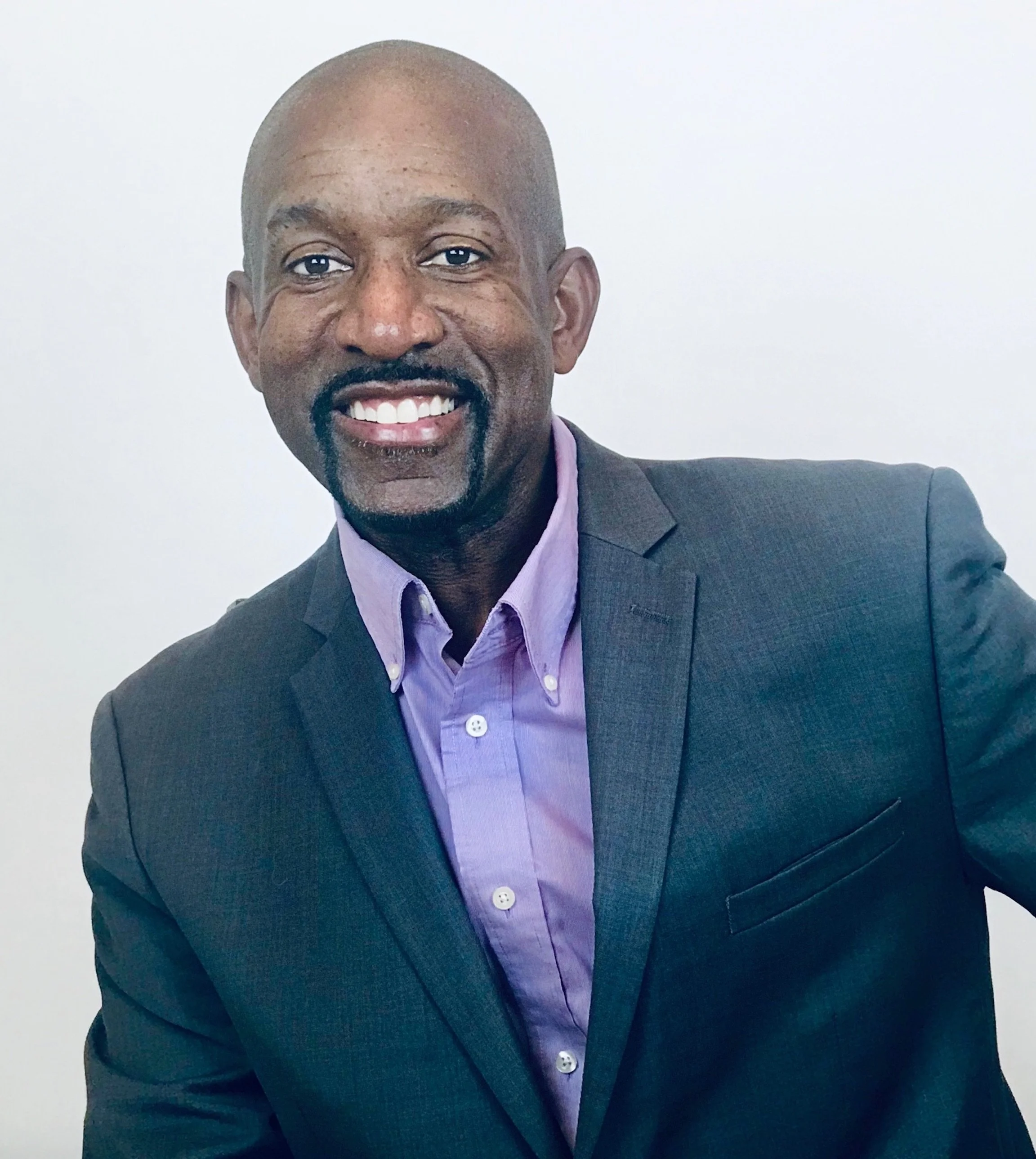 A smiling African American man with a bald head and a goatee, wearing a dark suit jacket and a purple dress shirt, standing against a plain white background.