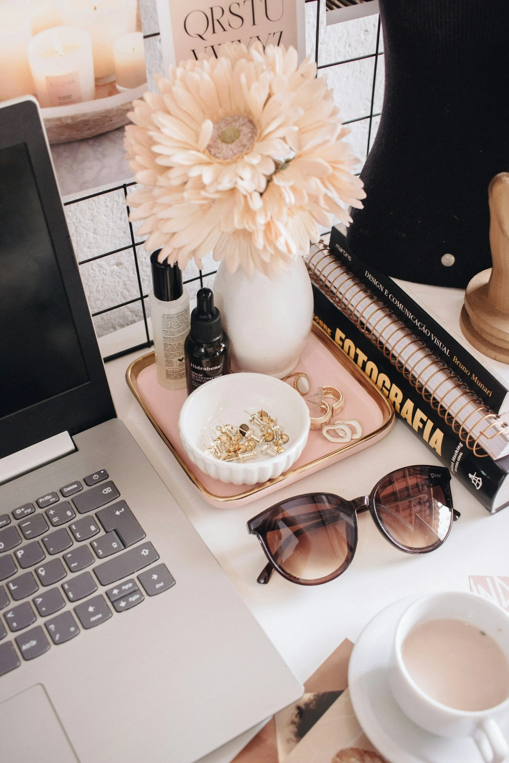 A workspace with a laptop, a pink tray holding a small white bowl with gold jewelry, a pair of sunglasses, various skincare bottles, a large sunflower in a white vase, and various books on a desk. There are candles in the background and a grid-style wall.