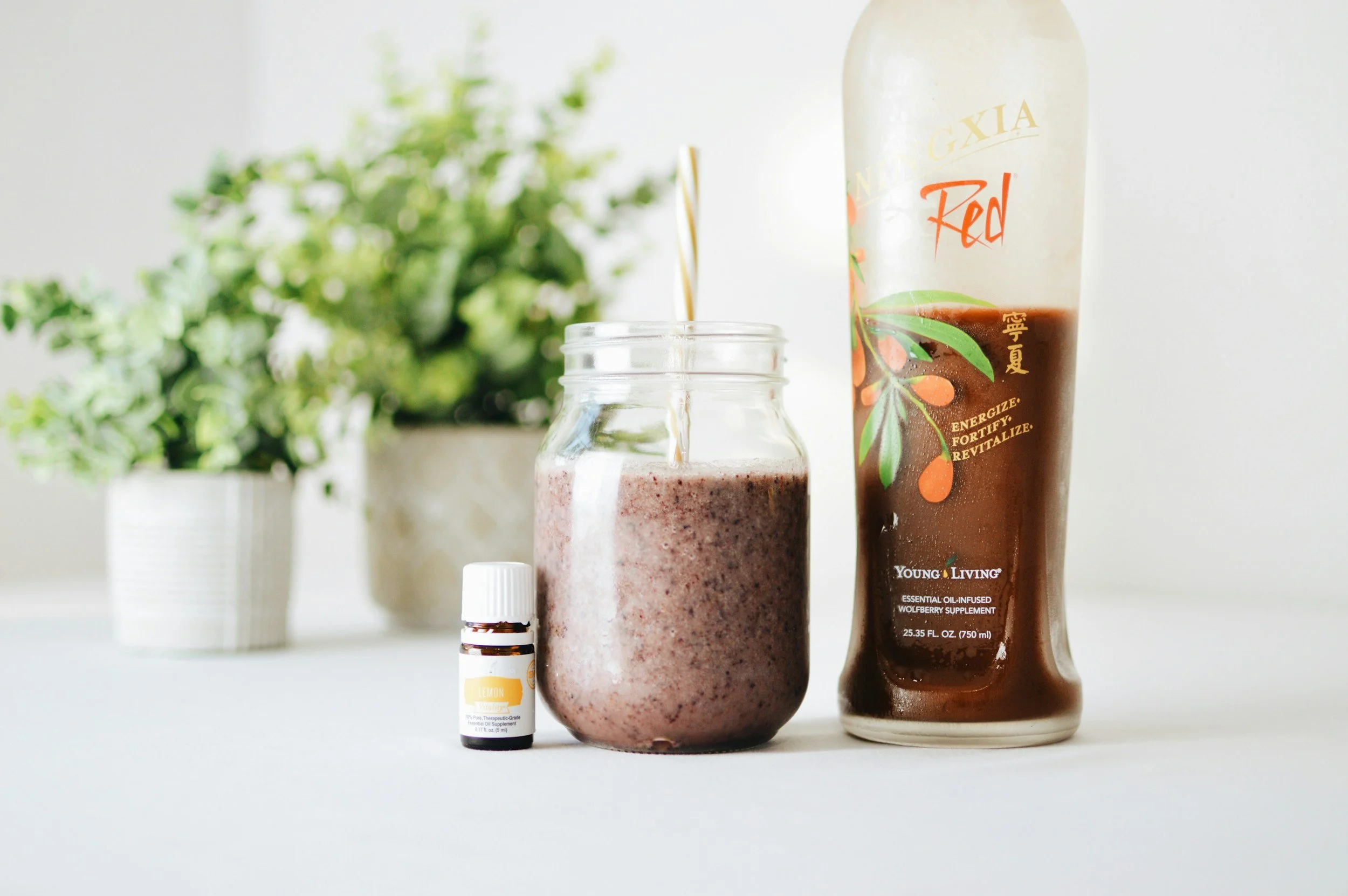 A glass jar filled with a pinkish smoothie, a small essential oil bottle, a tall bottle of berry supplement labeled Young Living with a floral design, and a potted plant in the background with green leaves.