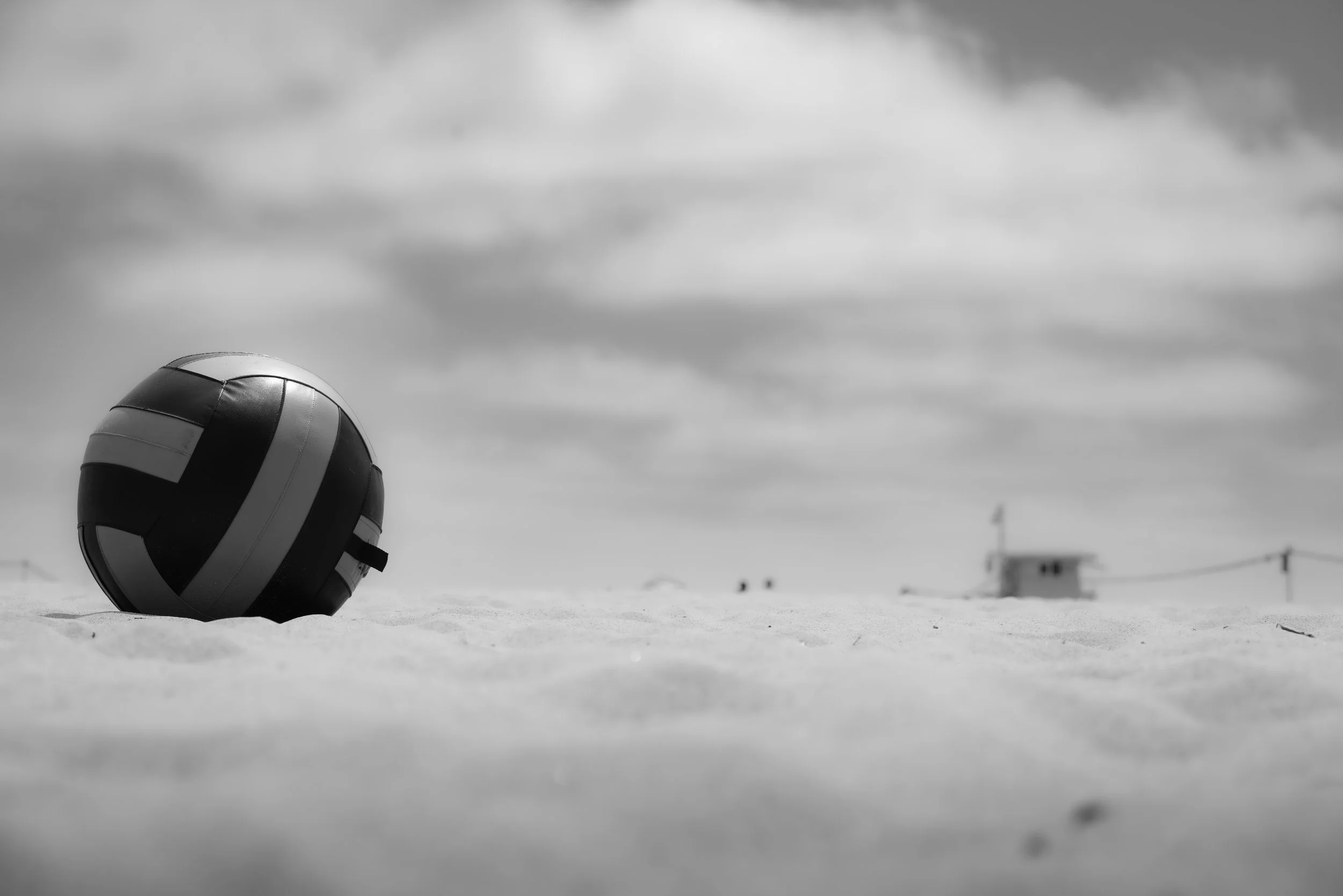 A black and white photograph of a volleyball resting on the sand at a beach or outdoor court, with a cloudy sky overhead.