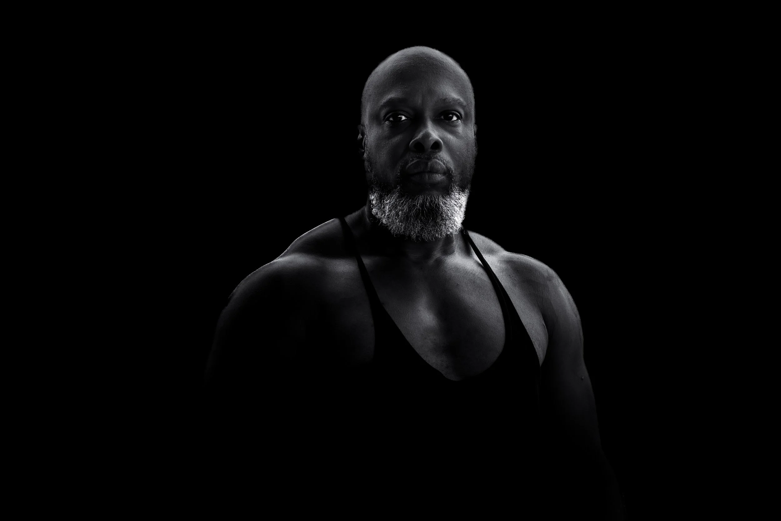 A portrait of a Black man with a gray beard, bald head, and wearing a black tank top against a black background.
