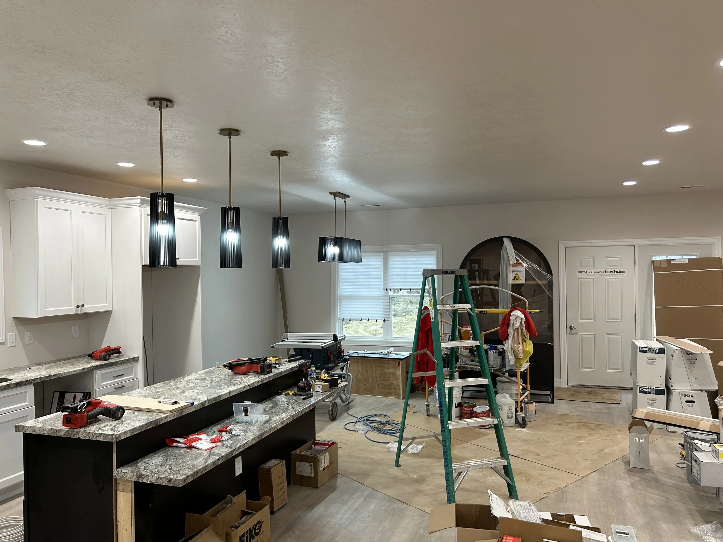 A kitchen undergoing renovation with tools, equipment, and construction materials. There are hanging pendant lights, a granite countertop island, cabinets, a ladder, and various boxes and supplies scattered around.