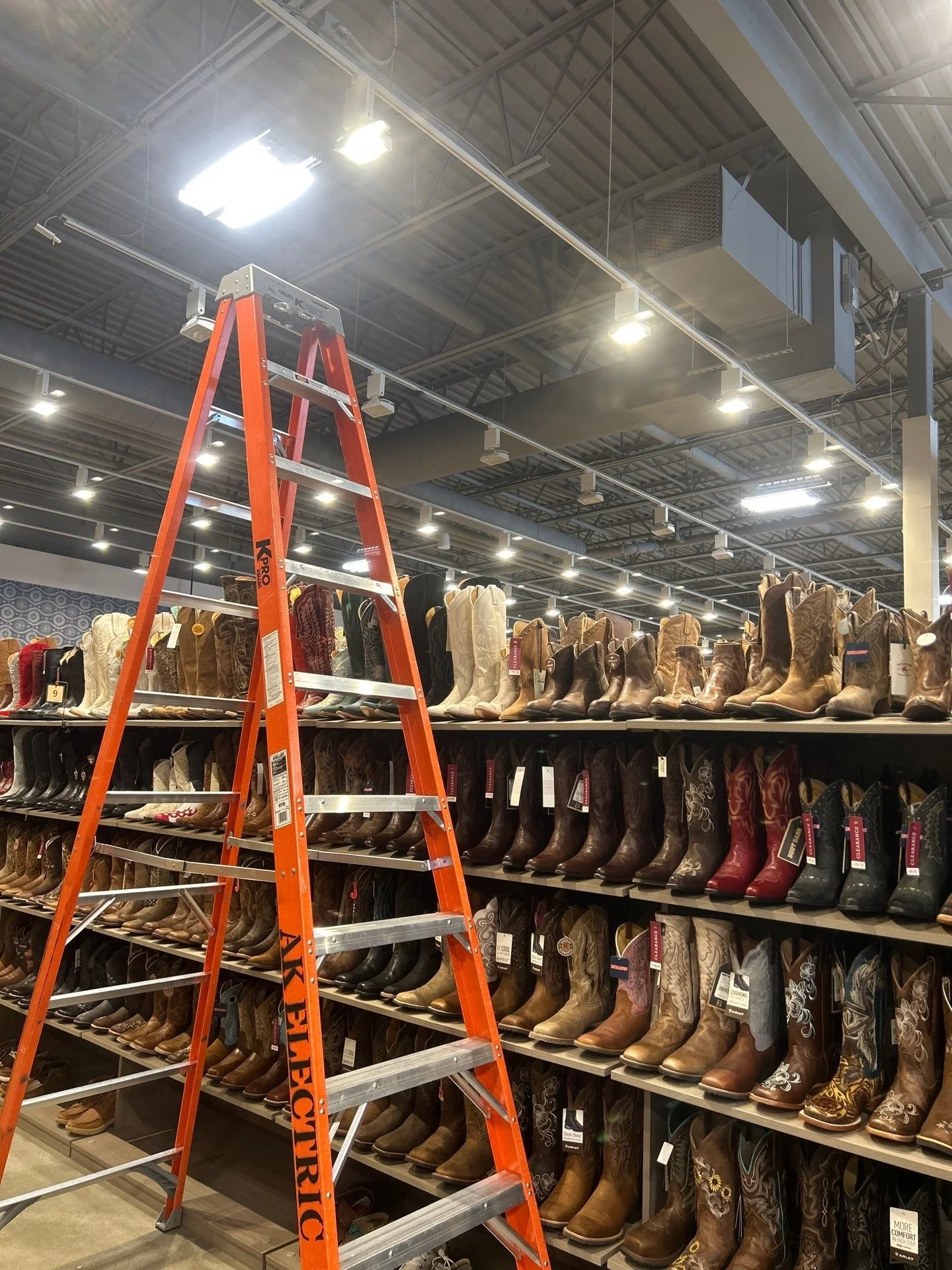 An orange Step ladder placed in front of shelves full of cowboy boots in a retail store. The store has a high ceiling with bright overhead lighting.