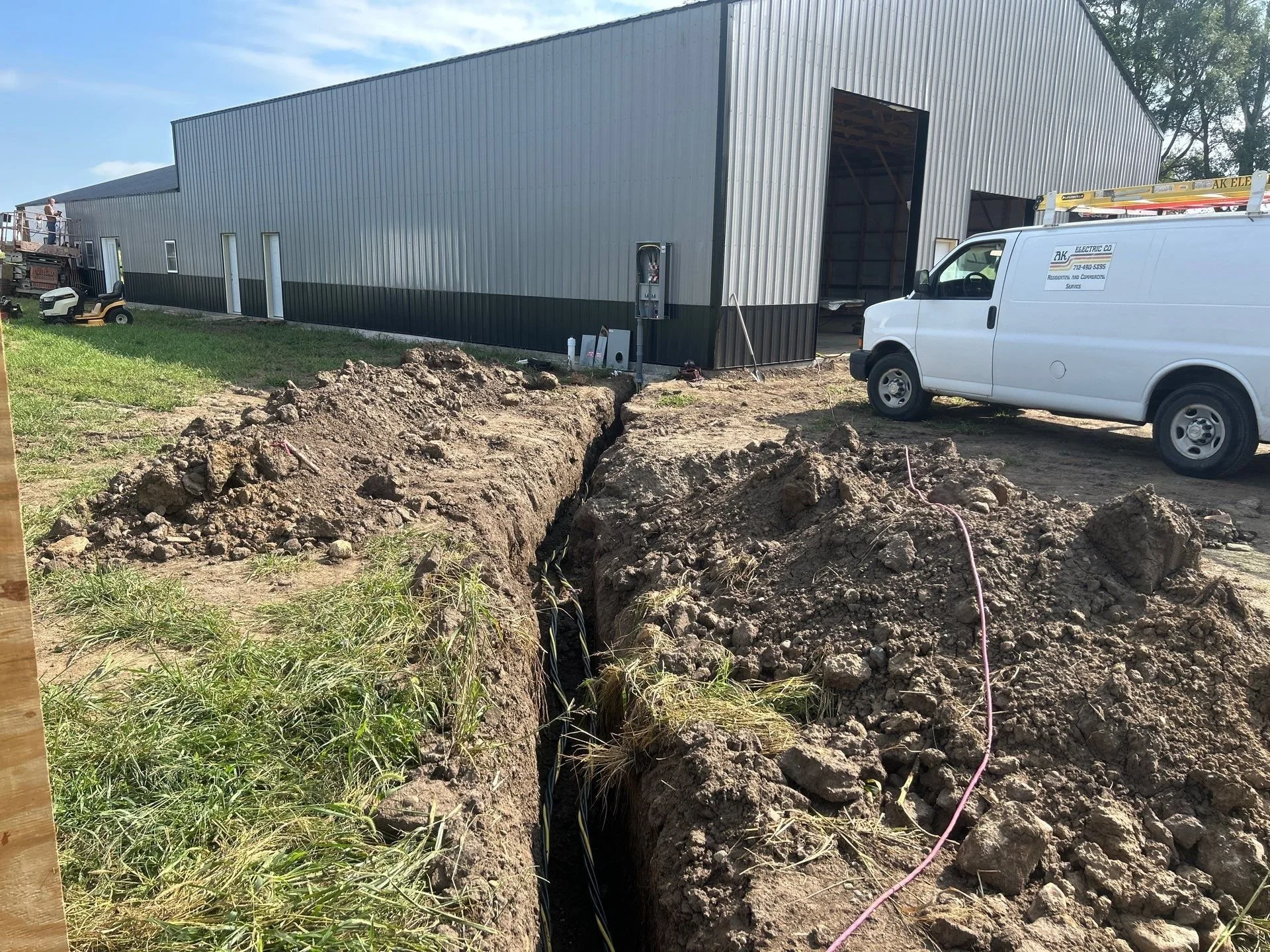 Construction site with a large metal building under construction, a white utility van with a ladder on top, and a deep excavation trench in the foreground.