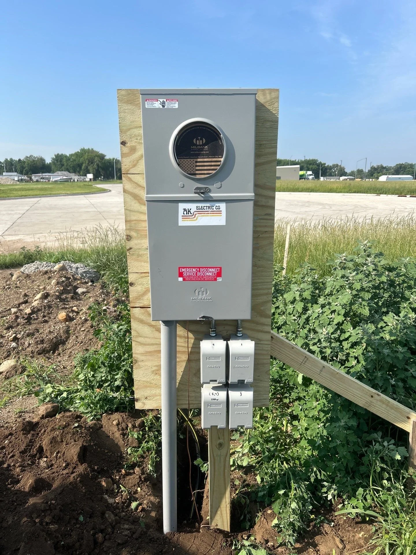 An temporary power installation with metal boxes and a round logo in a rural outdoor setting with grass and a road in the background.