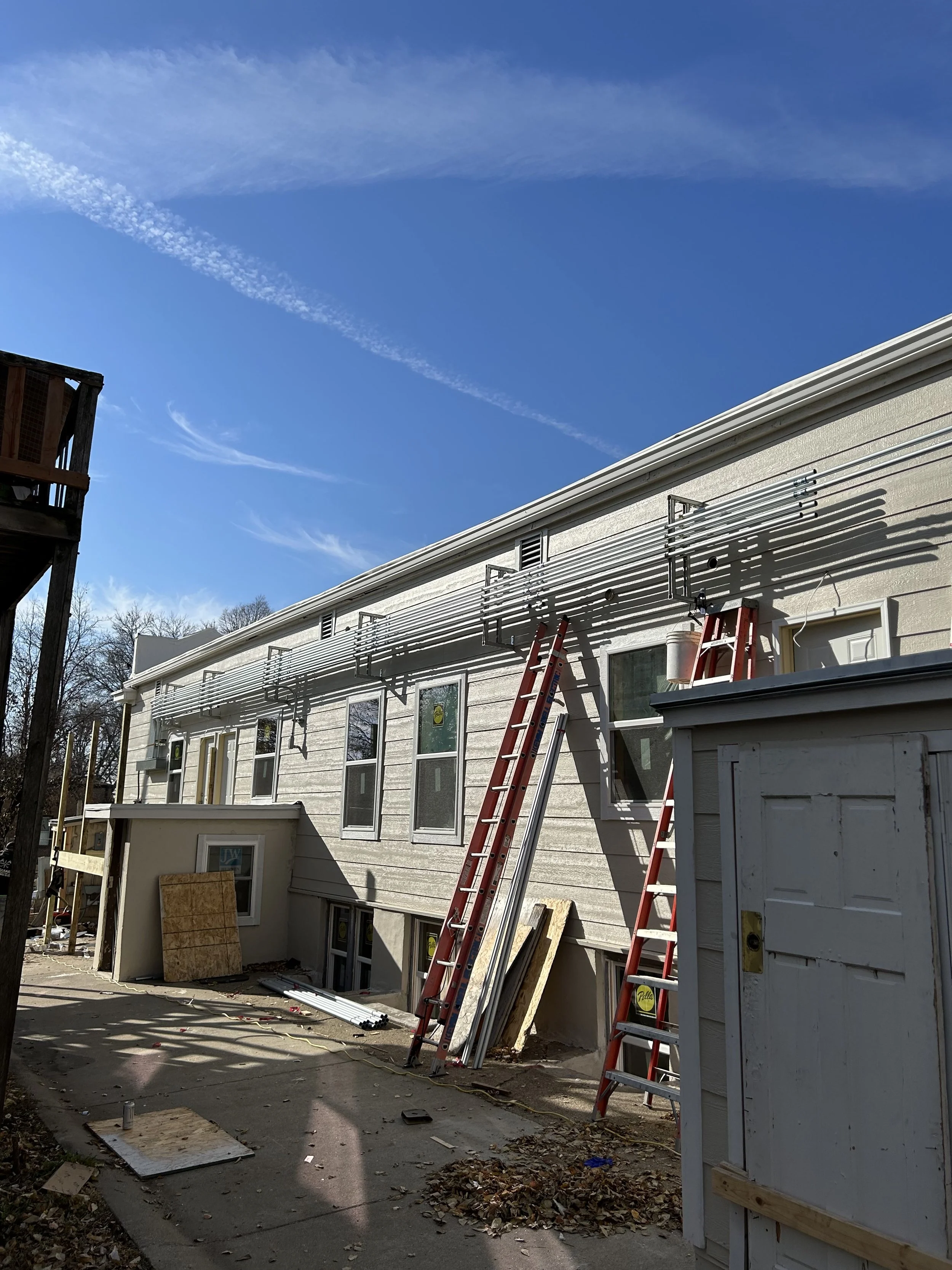 Construction site with ladders, building materials, and debris outside a residential building under a clear blue sky.