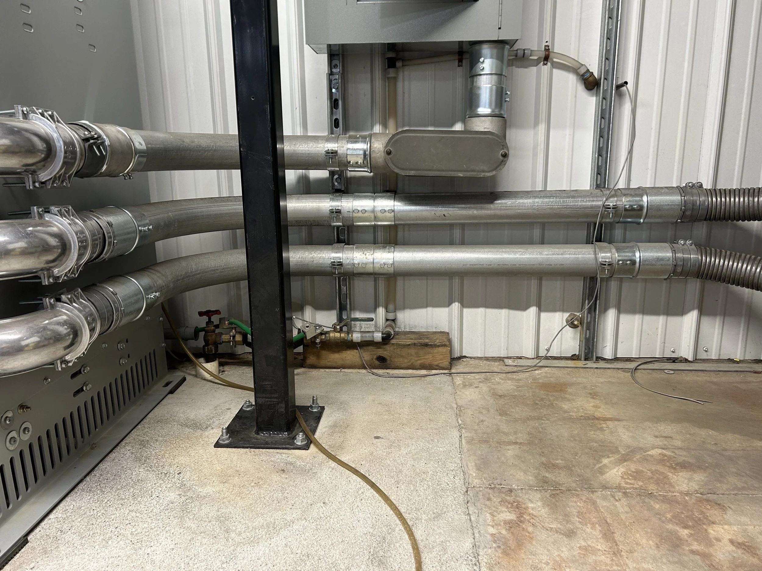 Industrial pipes and ductwork installed on an interior wall, secured with metal clamps and mounted on a concrete floor.