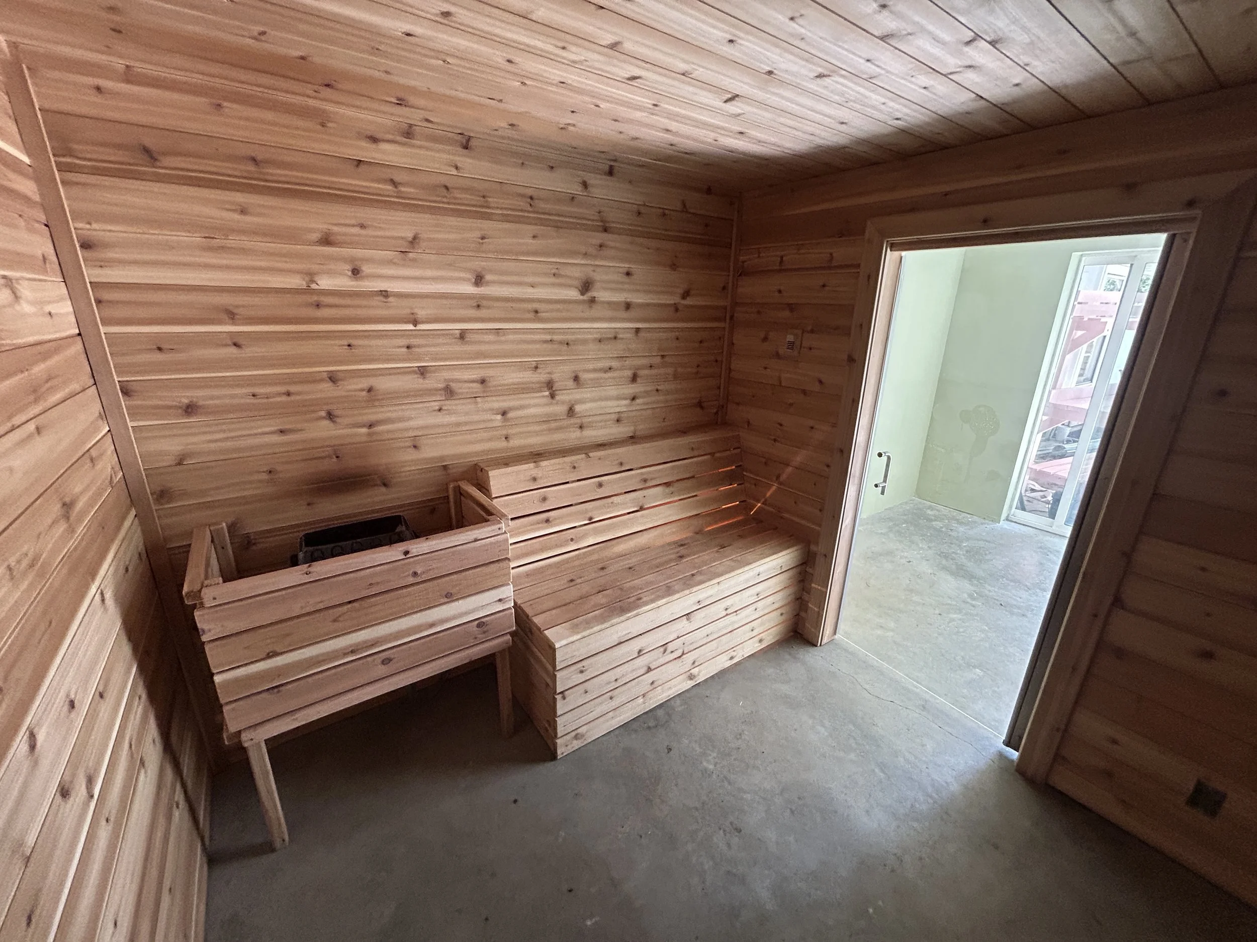 A room with wooden panel walls and ceiling, a wooden bench along the wall, and a glass door leading outside to a balcony or patio.