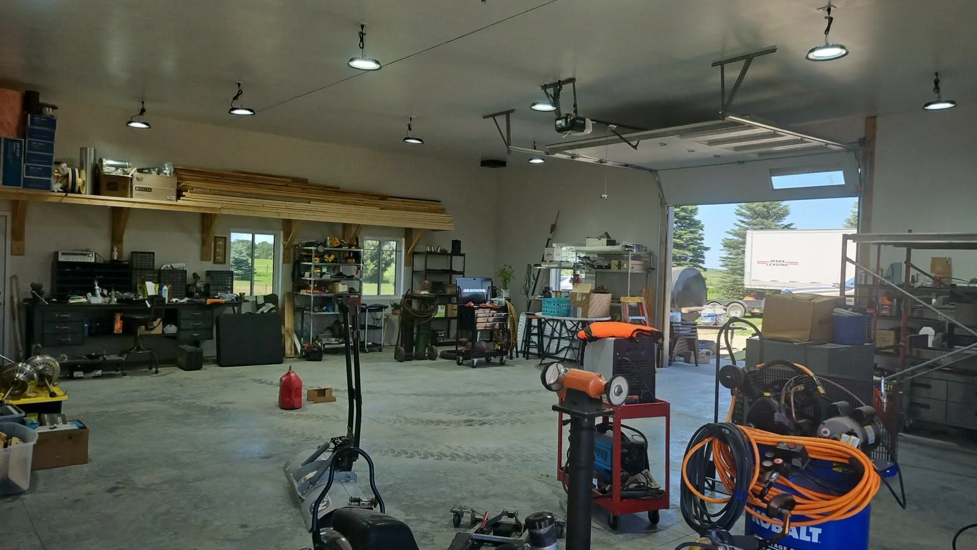 A spacious workshop or garage with various tools, equipment, and supplies. The area has unfinished walls, a concrete floor, and a garage door opening to the outside where trees and a truck are visible. Items include workbenches, shelves, power tools, extension cords, and a small portable heater.