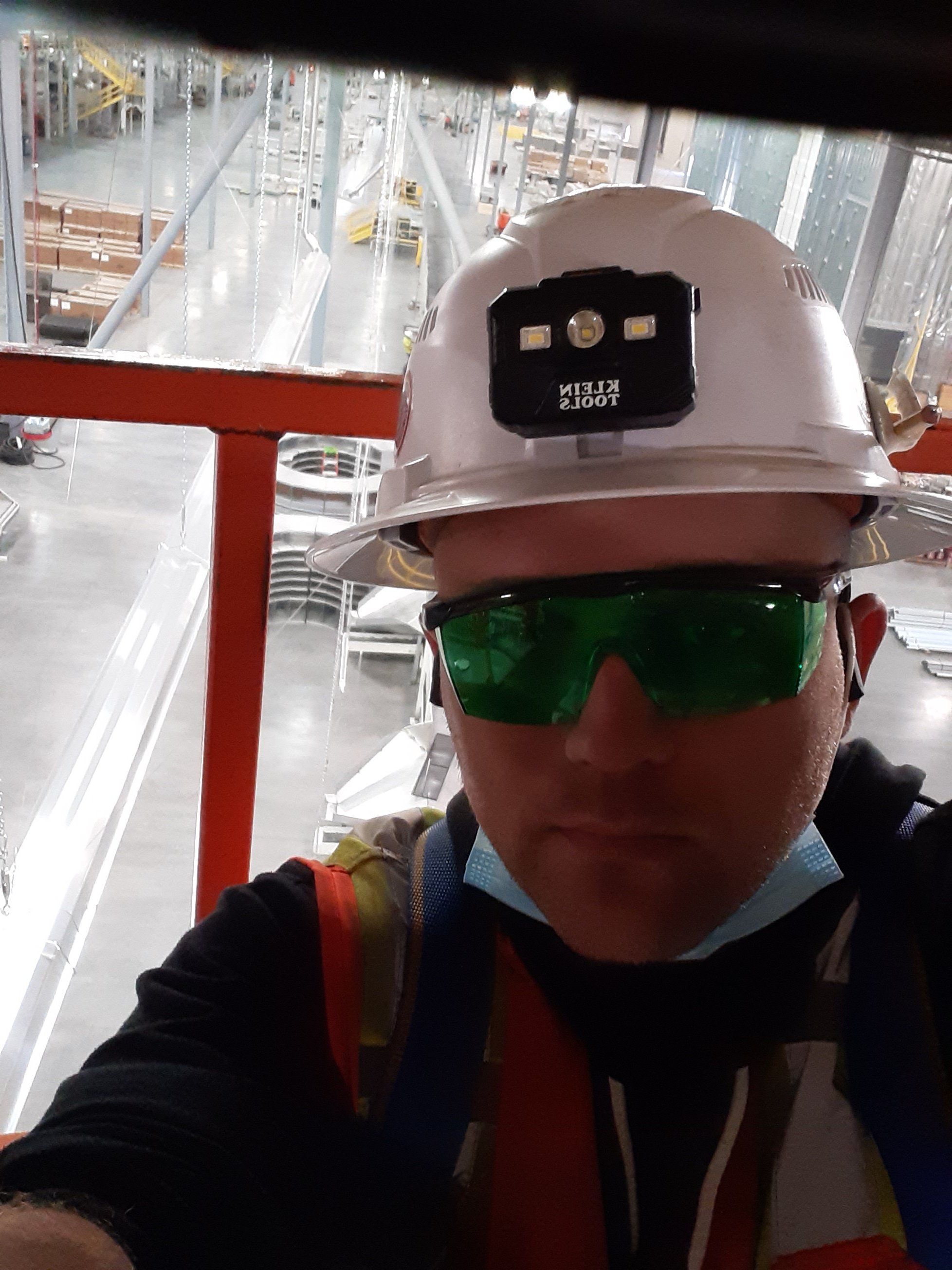 Worker wearing a white hard hat with a mounted LED headlamp, green safety glasses, and a black jacket with a red strap, taking a selfie inside an industrial warehouse.