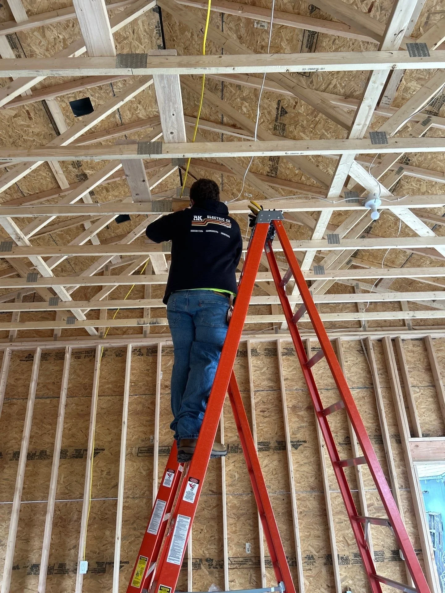 A person working on electrical inside a building under construction, standing on an orange ladder, with exposed wooden beams and OSB walls.