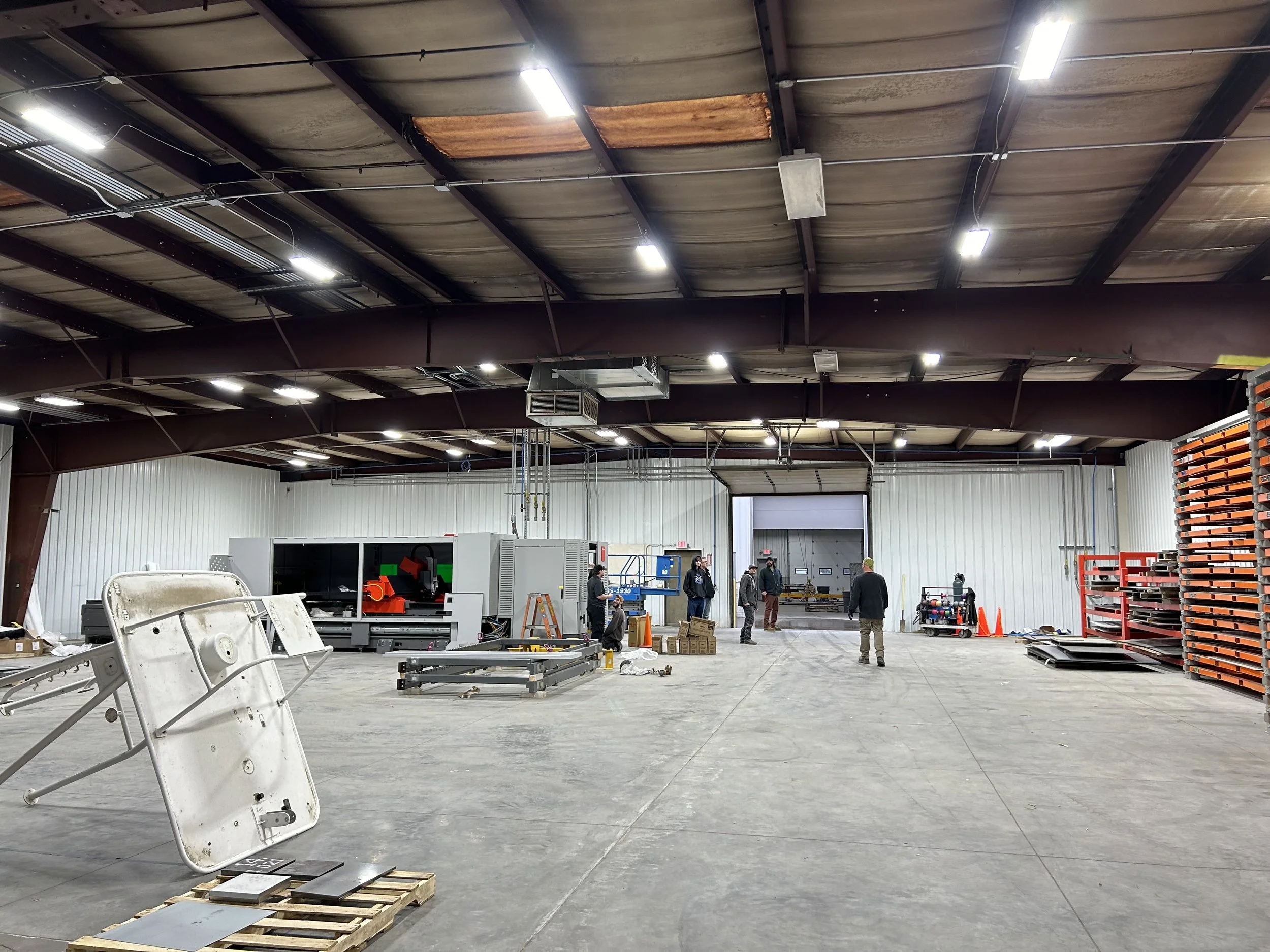 Inside a large industrial warehouse with a high ceiling made of metal panels. Several workers are scattered across the space, engaged in various tasks. There are various pieces of equipment, shelves, and materials, including a large machine, pallets, and a disassembled bed frame in the foreground. Bright ceiling lights illuminate the space.