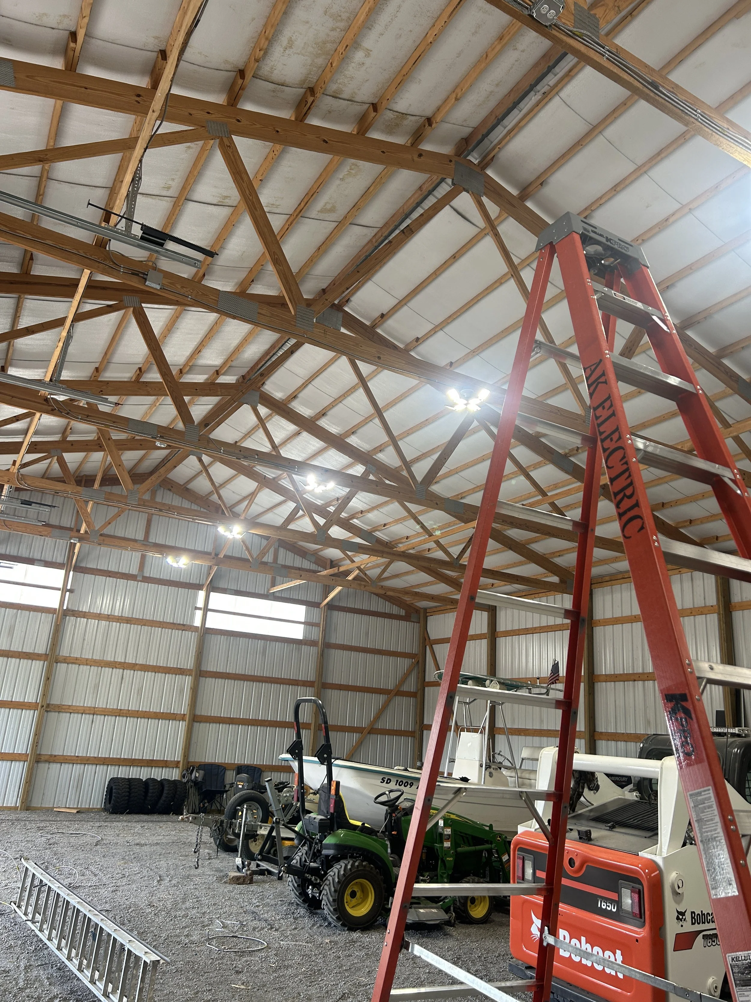 Inside a large garage or workshop with unfinished wooden rafters and metal walls, containing various tools, a boat, a small green tractor, tires, a red ladder, and industrial lighting.