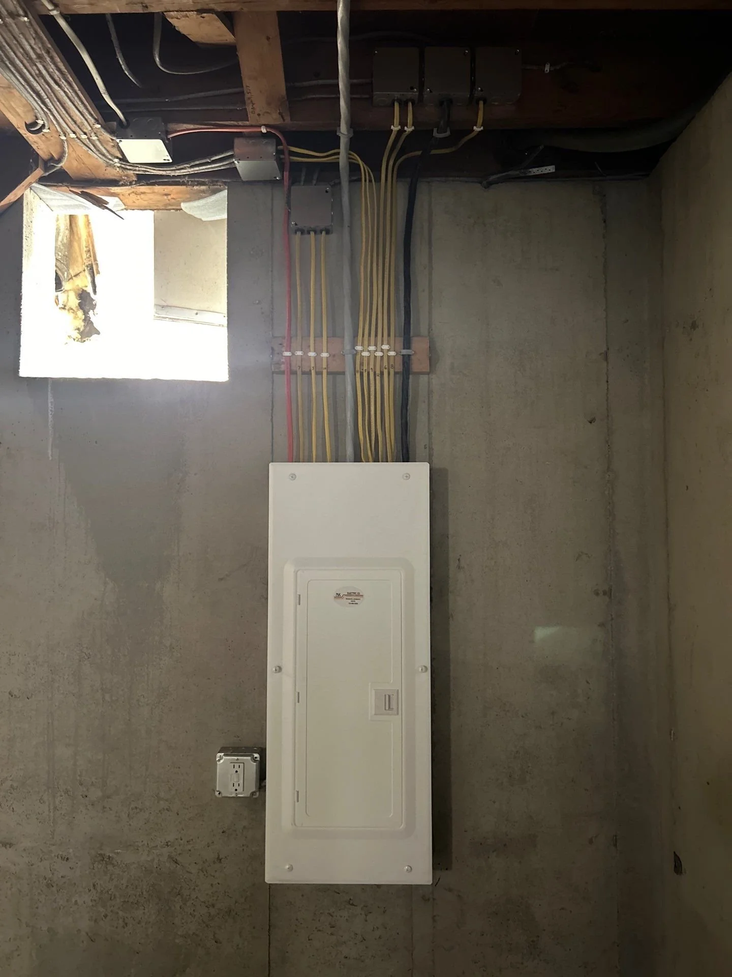 Electrical panel on a concrete wall with colored wires connected and an open window with damaged wooden frame above.