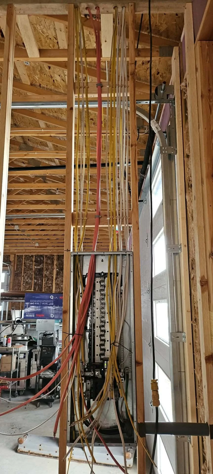 View of a construction site with exposed wooden framing and electrical wiring, including red, yellow, and white cables, with a garage door and some tools in the background.