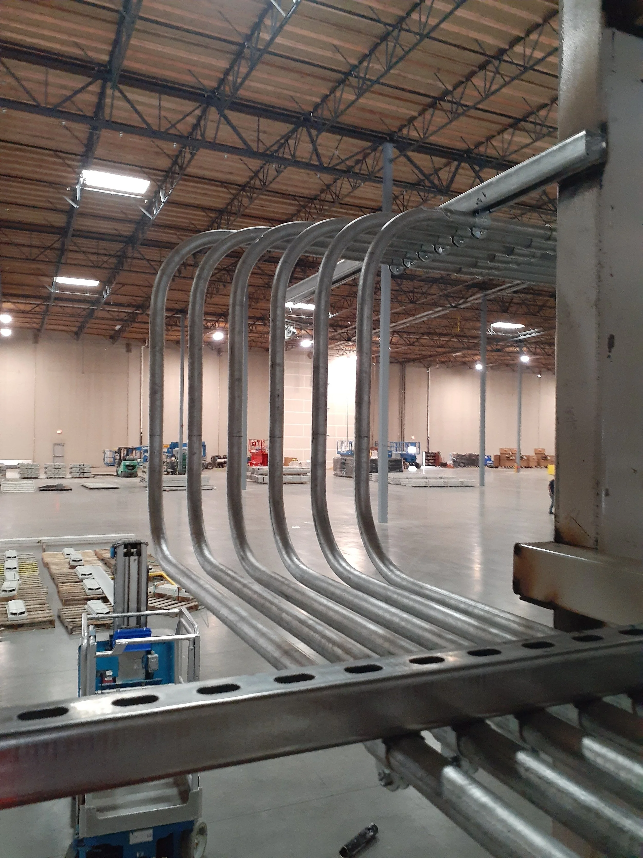 Inside a large warehouse under construction, metal pipes are organized in bundles and railings. The ceiling features exposed trusses and installed lighting, with various construction equipment and materials in the background.