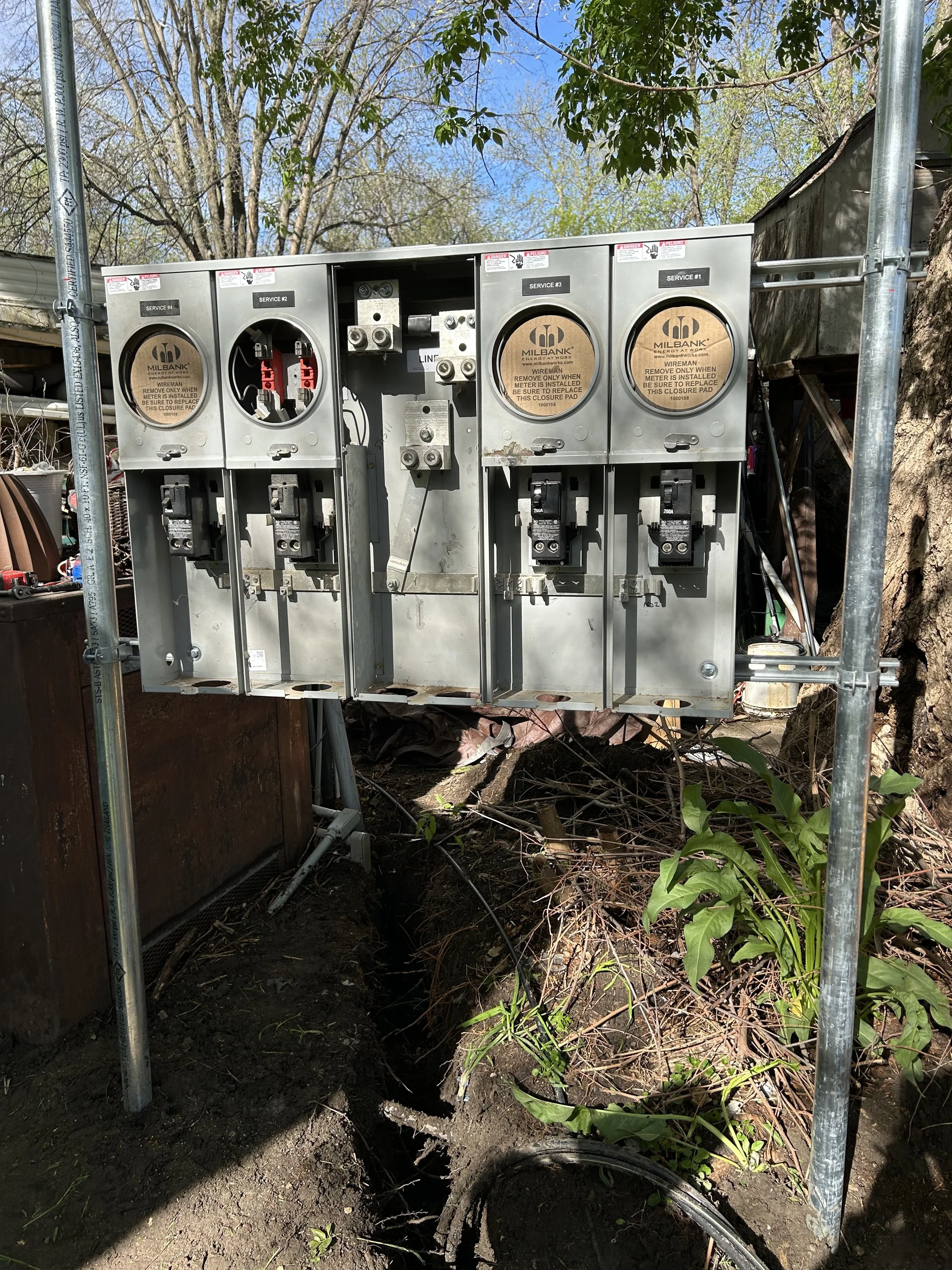 Electrical meter box installed outdoors with wires and labeled compartments, surrounded by trees and greenery.