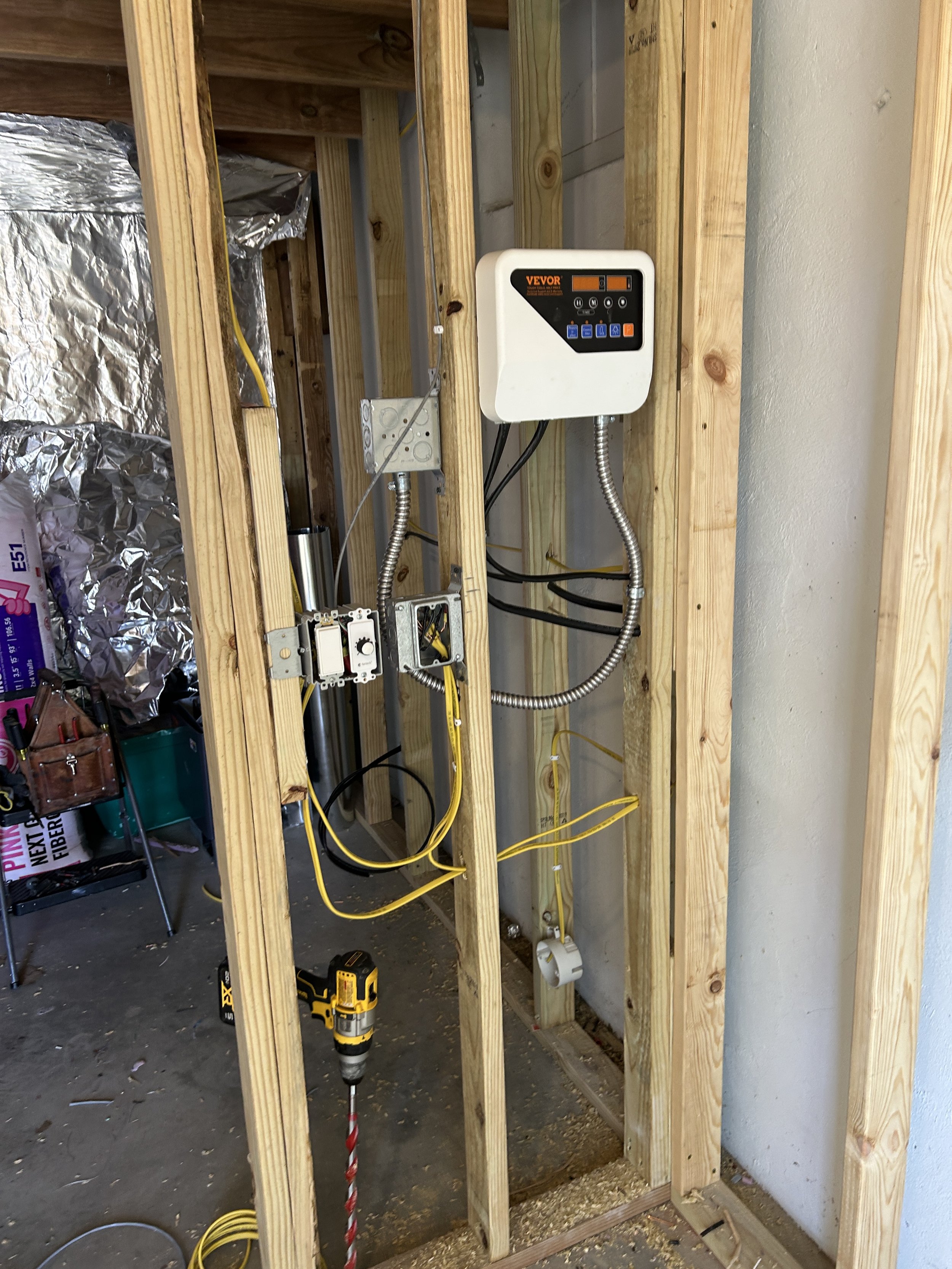 A building interior framing with electrical wiring, a white electrical control box labeled VEOR, metal conduits, and some tools on the floor.