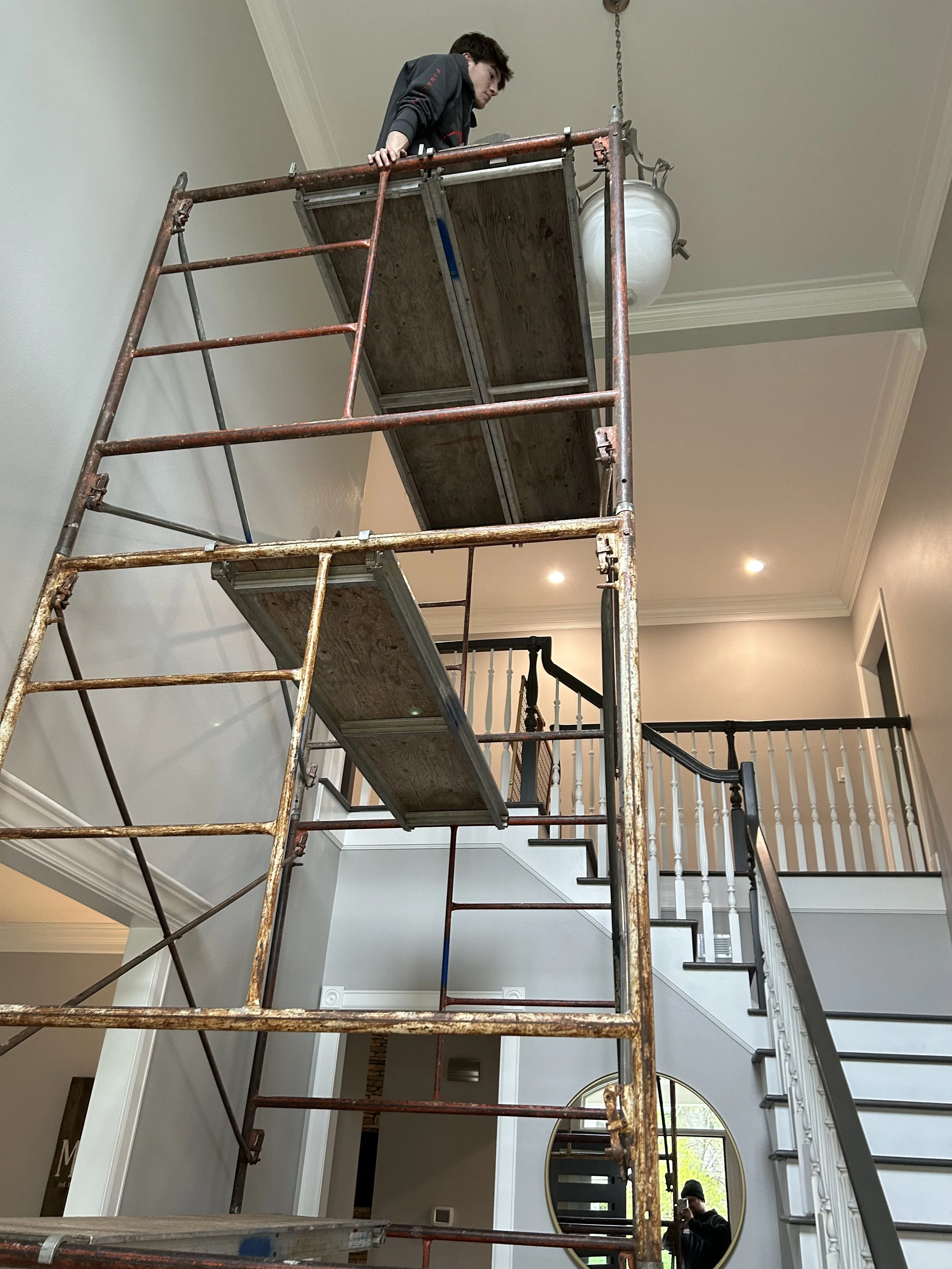 A person standing on scaffolding inside a house, working on the ceiling near a chandelier, with a staircase and a mirror reflecting a person taking the photo.