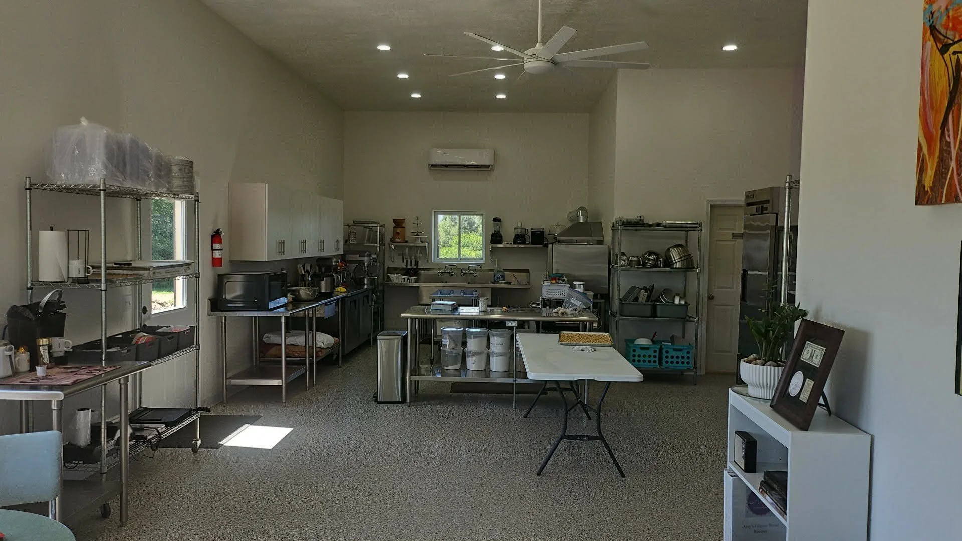 A kitchen or culinary workspace with metal shelves, stainless steel counters, cooking utensils, and appliances, illuminated by recessed ceiling lights and natural light from windows.