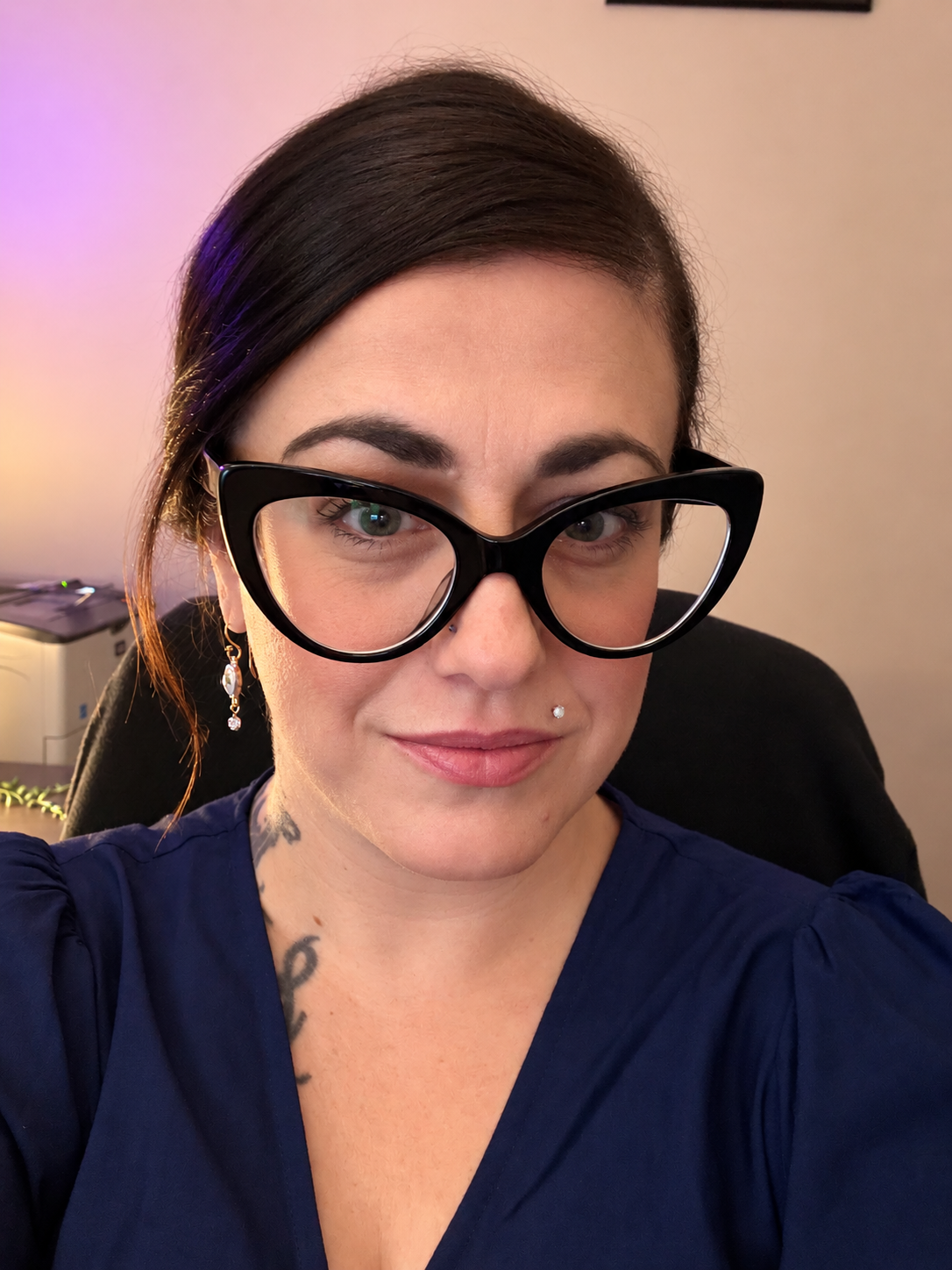 Woman with short dark hair and large black glasses taking a selfie in an office with a computer and colorful lighting in the background.