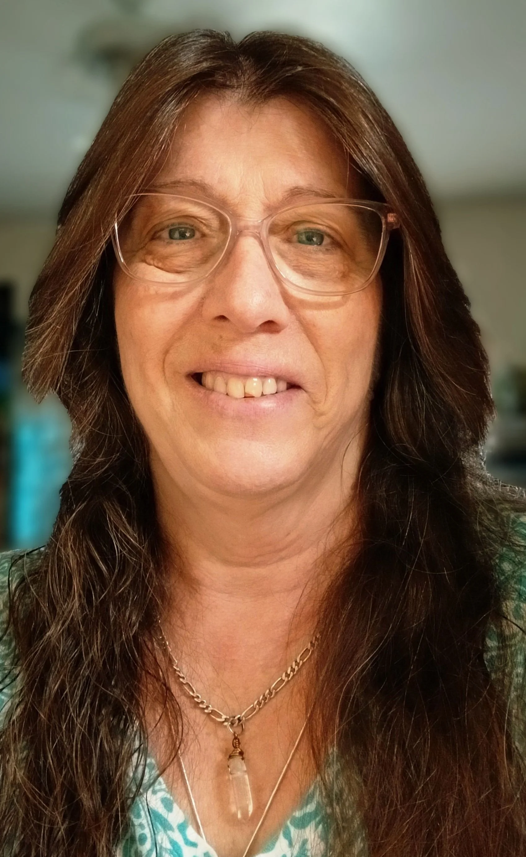 Close-up of a woman with long brown hair, wearing glasses, gold jewelry, and a patterned top, smiling inside a room.