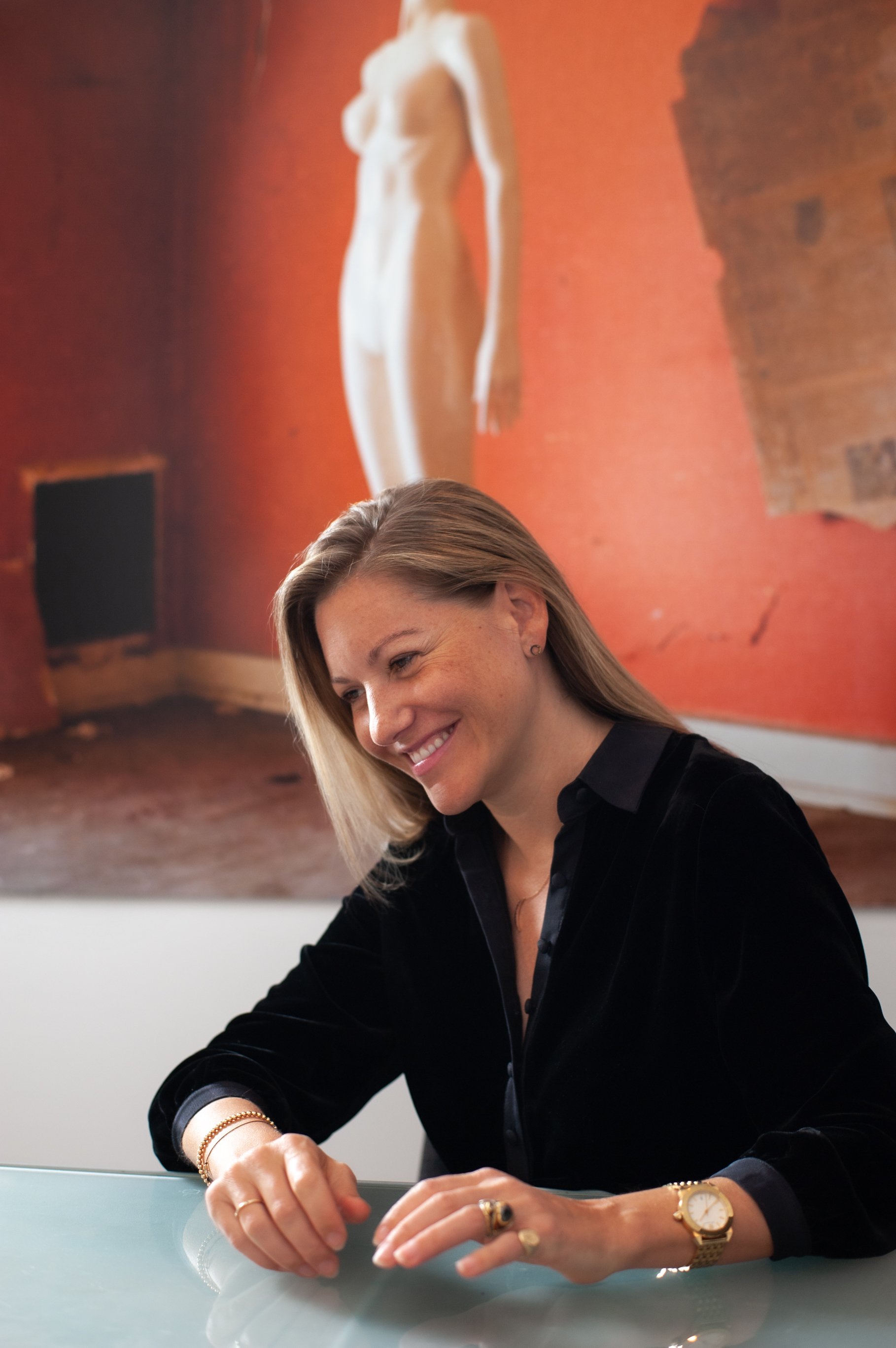 A woman with blonde hair smiling, wearing a black velvet jacket and various jewelry, sitting at a reflective table with a large painting of a nude woman and a brick building behind her.
