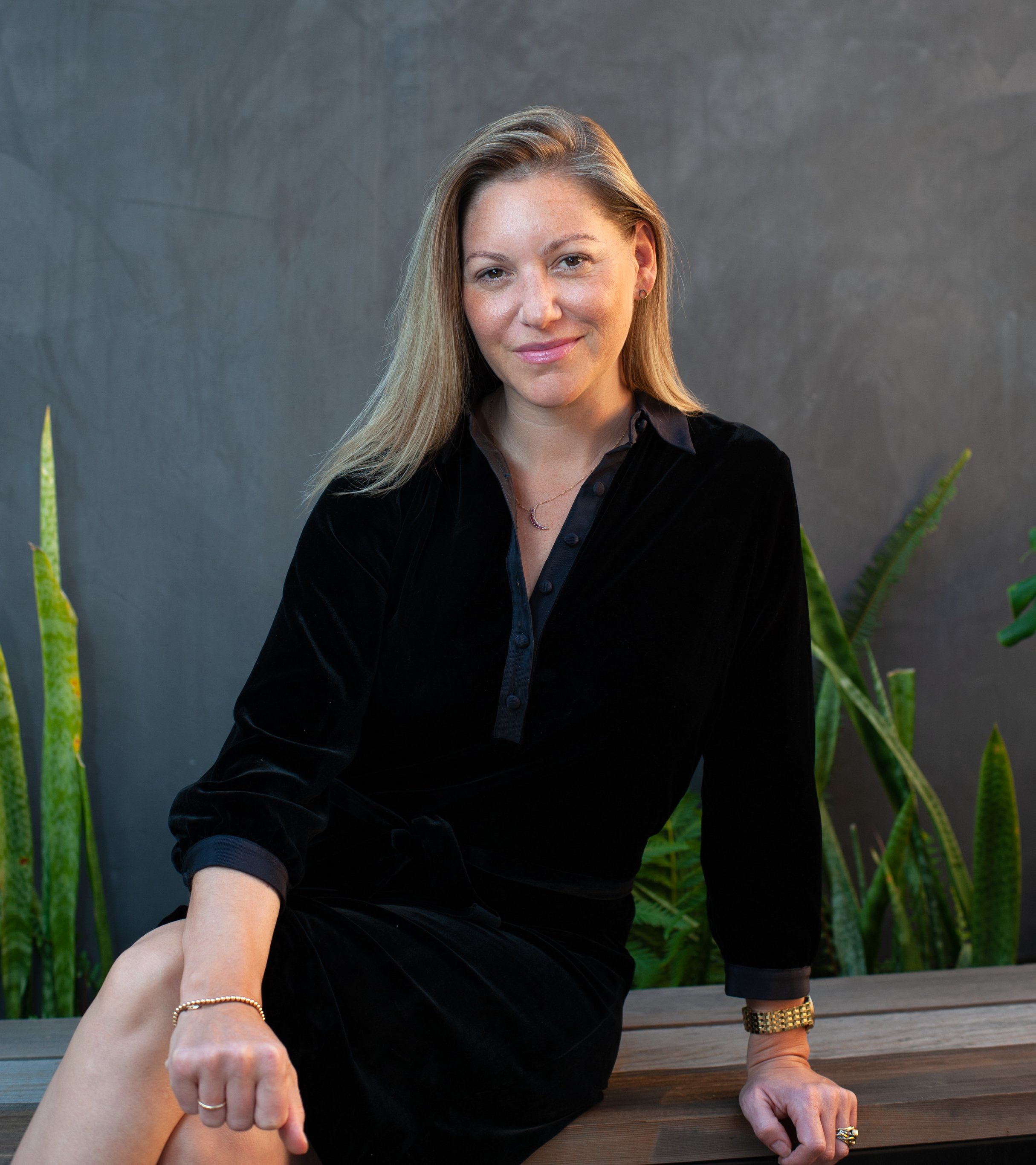A woman with long blonde hair, wearing a black shirt and gold jewelry, sitting on a wooden bench outdoors surrounded by green plants.