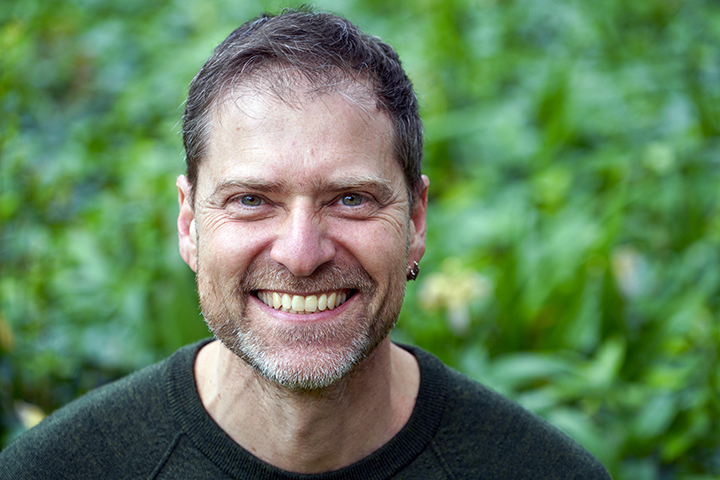 A smiling man with short brown hair, a beard, and a mustache, wearing a dark sweater, standing outdoors with green foliage in the background.