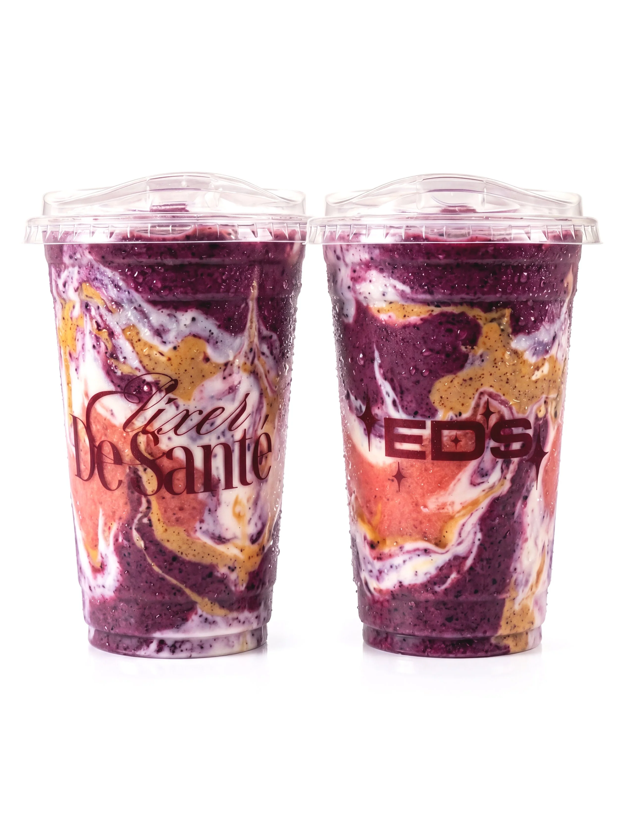 Two beautifully crafted berry swirl smoothies served in clear cups with dome lids, showcasing vibrant layers of rich berry colors and creamy textures. Each cup features the Elixer De Sante branding, creating a refreshing and eye-catching presentation
