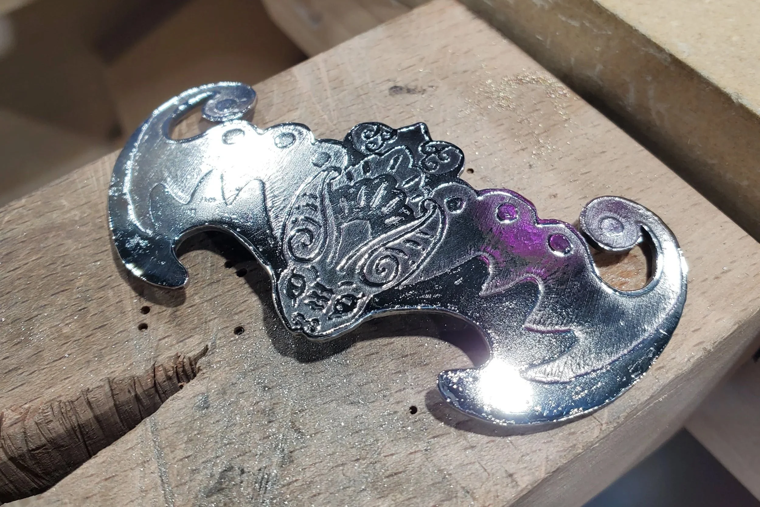 Close-up of a bat metal pendant with intricate swirling patterns placed on a wooden work surface.