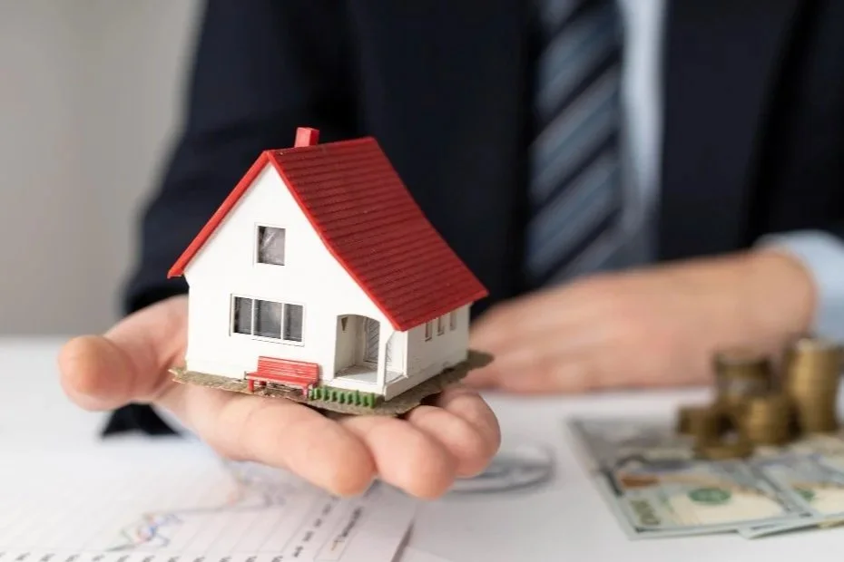Person in business attire holding a small model house with a red roof, with stacks of coins and money on a desk in the background.