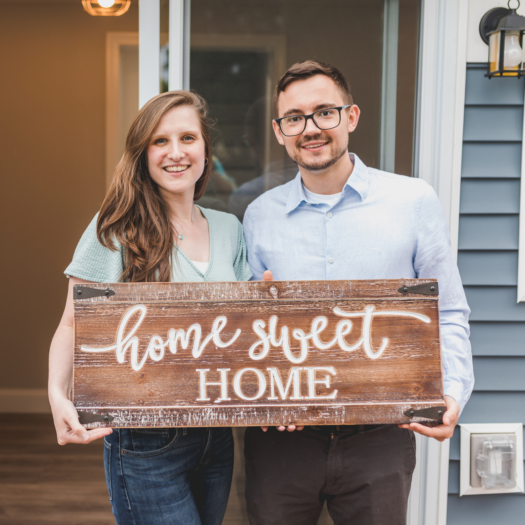 A smiling couple holding a wooden home decor sign that reads 'home sweet home' in front of their house.