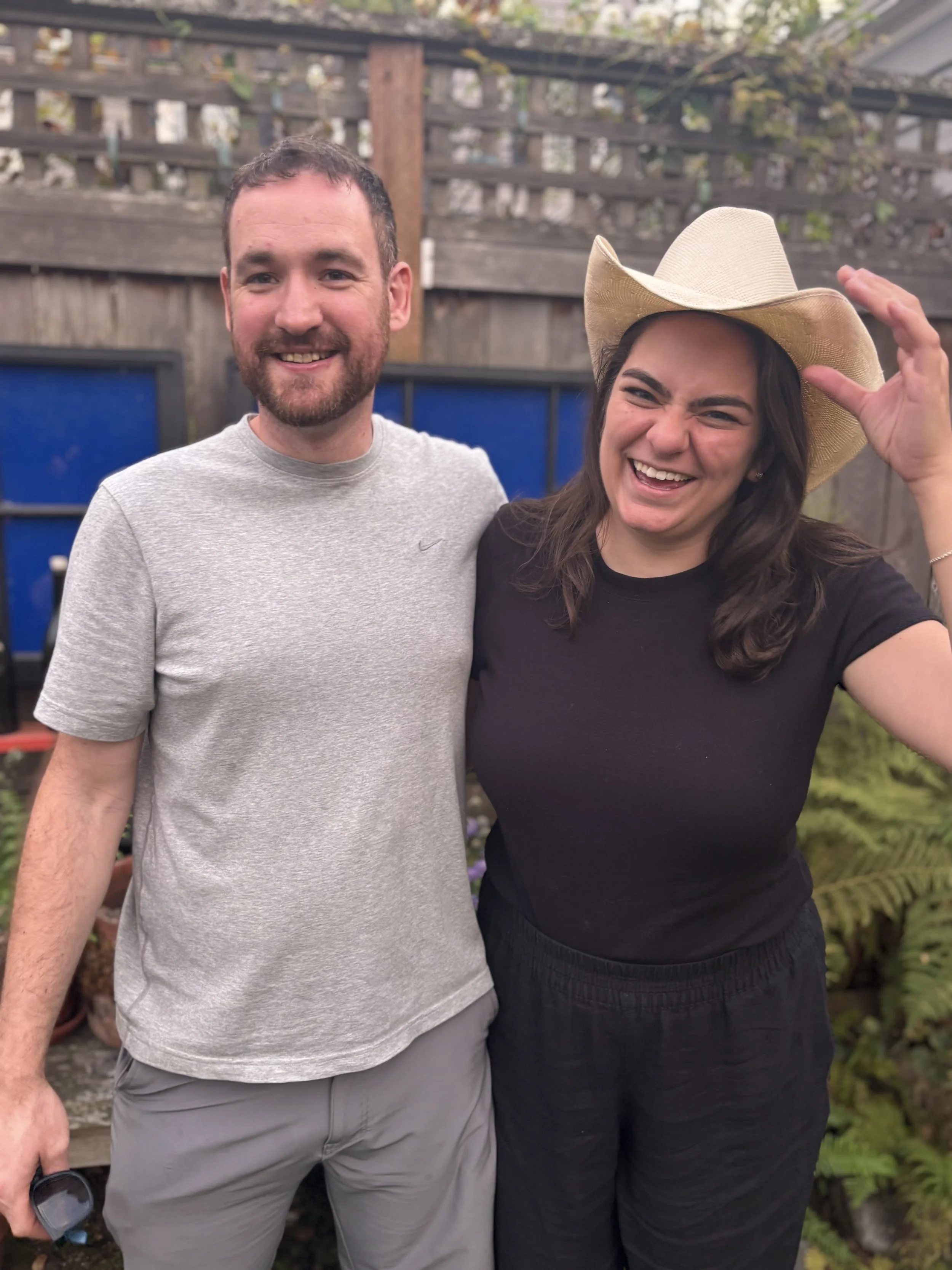 A man and woman standing together outdoors, smiling, with a wooden fence, plants, and gardening supplies in the background.