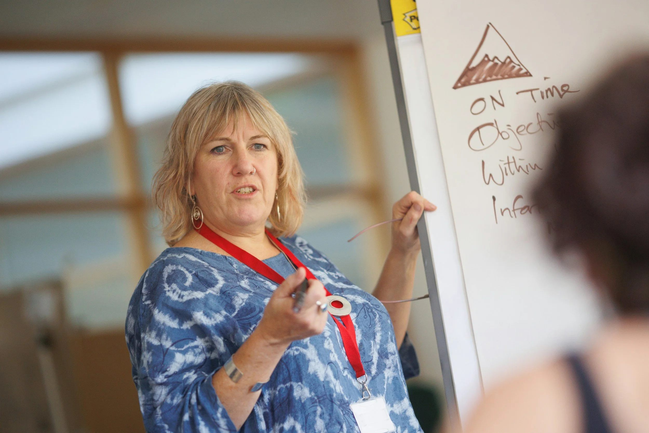 A woman with blonde hair, wearing earrings and a blue patterned shirt, pointing at a whiteboard with writing on it. The whiteboard has a drawing of a mountain and the text "On Time" visible. The background is blurred.