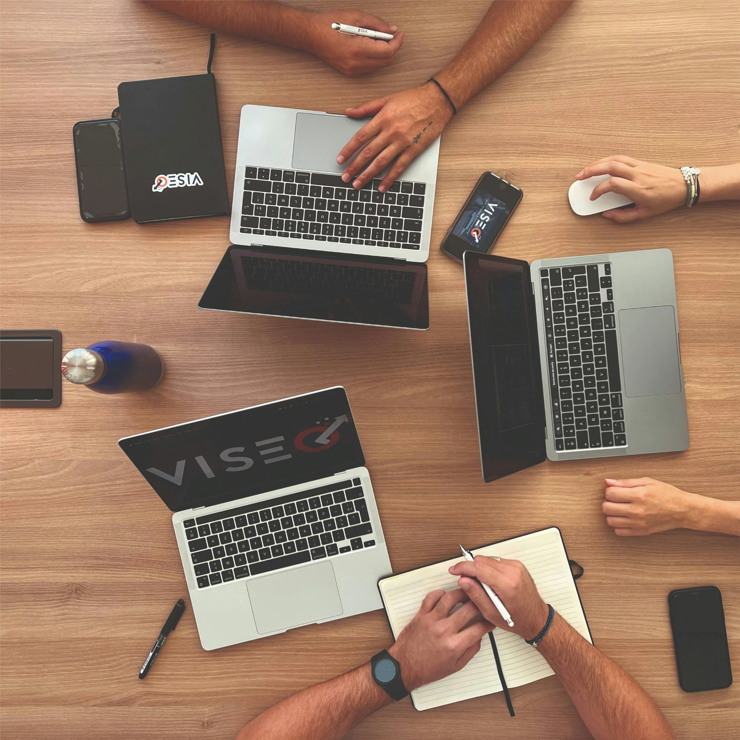 Overview of a work desk with multiple electronic devices, notebooks, pens, and hands of two people working together.