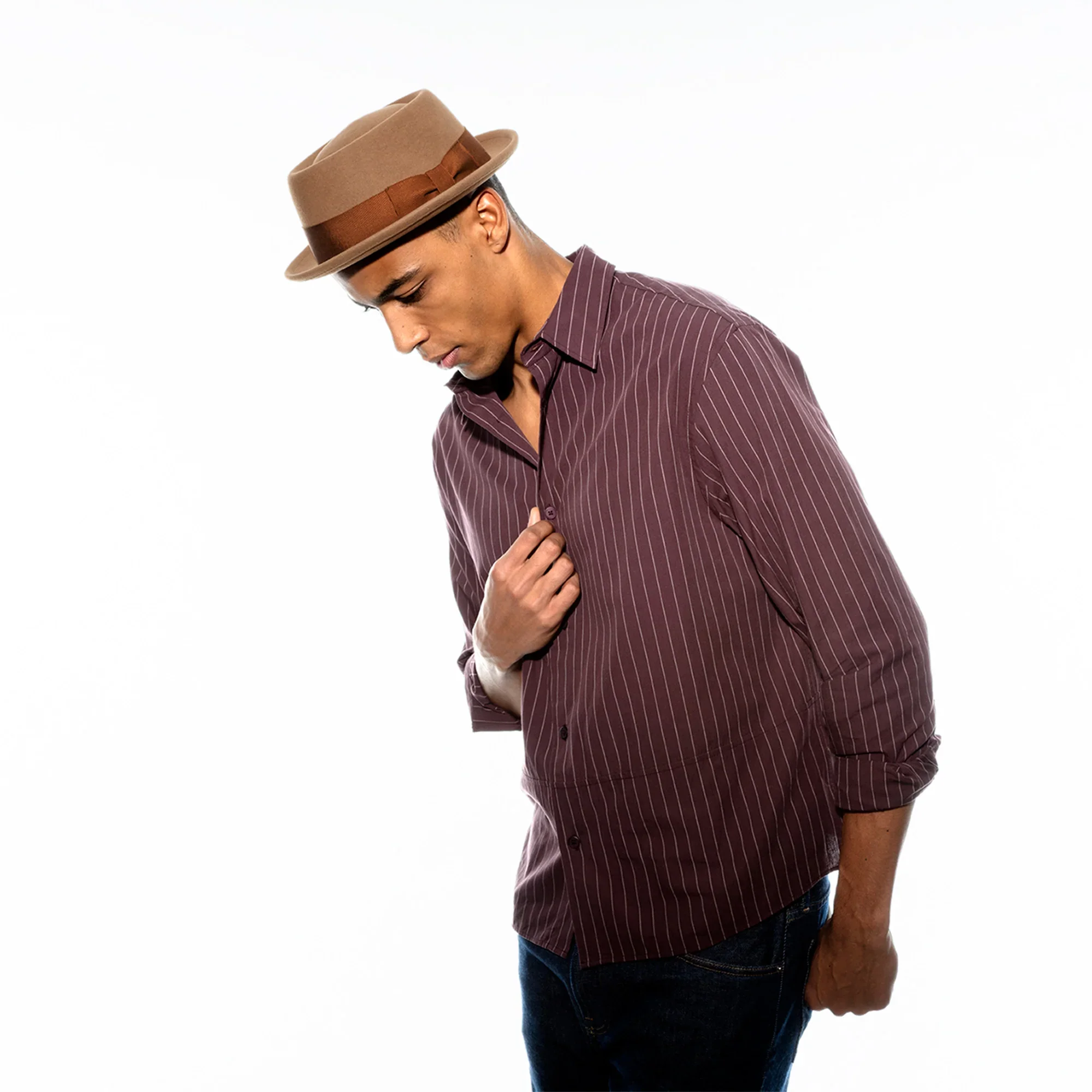 A young man wearing a brown fedora hat and a maroon pinstriped shirt stands against a white background. He is looking downward with his eyes closed and has his hand on his chest.