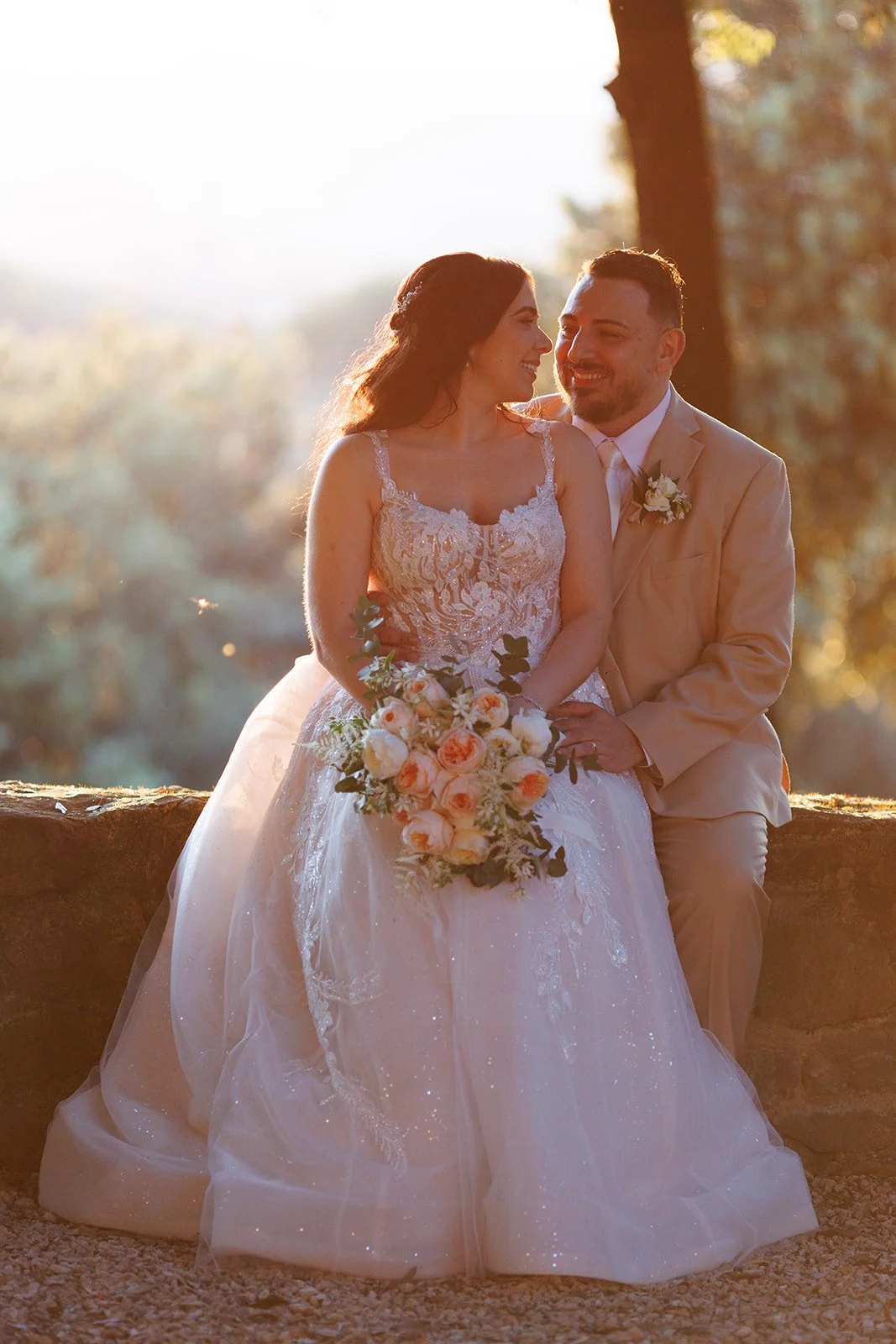 A bride and groom sitting close together outdoors at sunset, smiling at each other. The bride is holding a bouquet of peach and white roses, wearing a lace wedding dress. The groom is in a beige suit with a boutonniere. There are trees and sunlight i