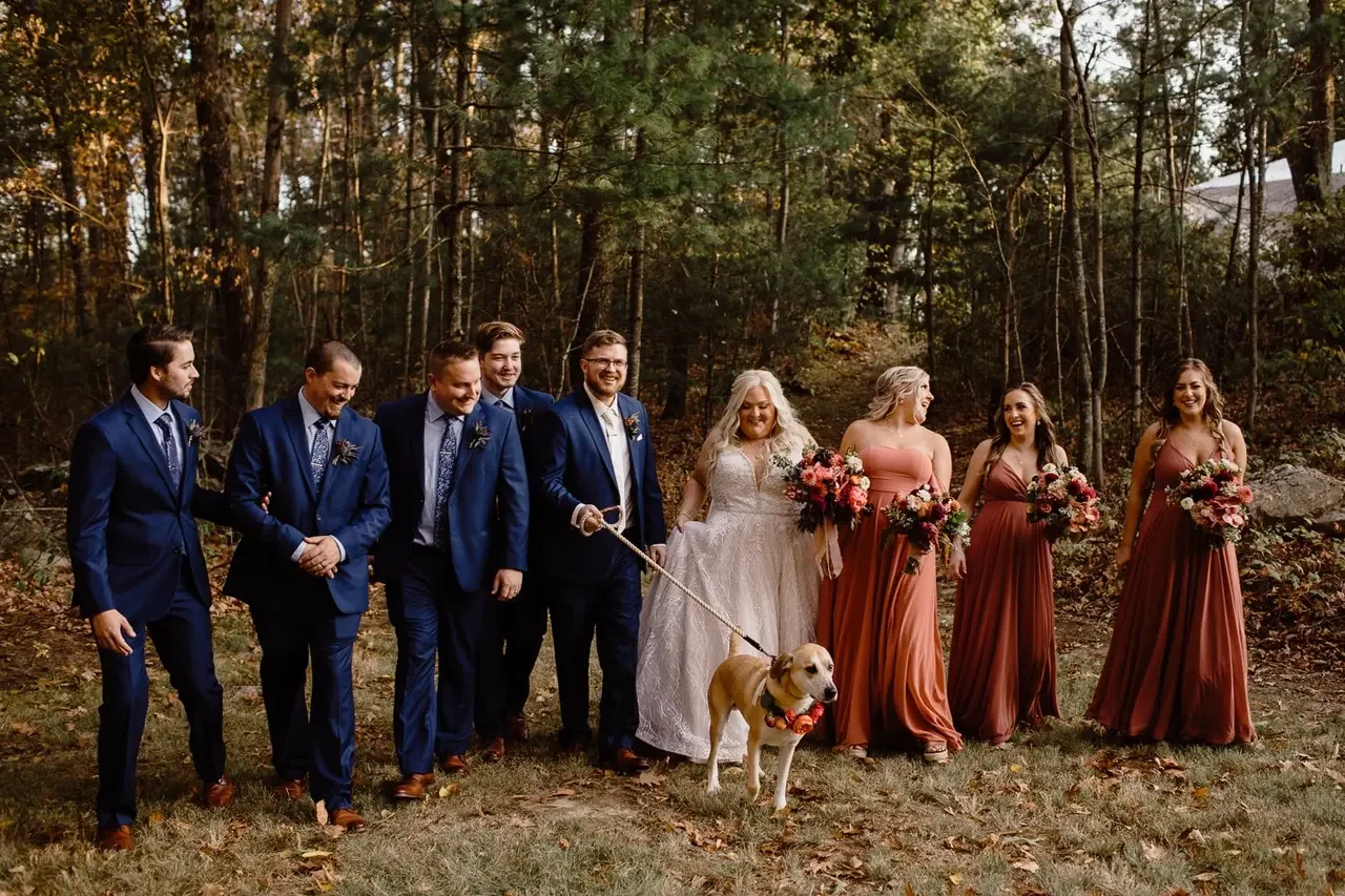 A wedding party outdoors with the bride and groom, groomsmen, and bridesmaids standing on grass. The bride is holding a bouquet and a tan dog with a flower collar; the bridesmaids are holding bouquets and are in rust-colored dresses. The groomsmen ar