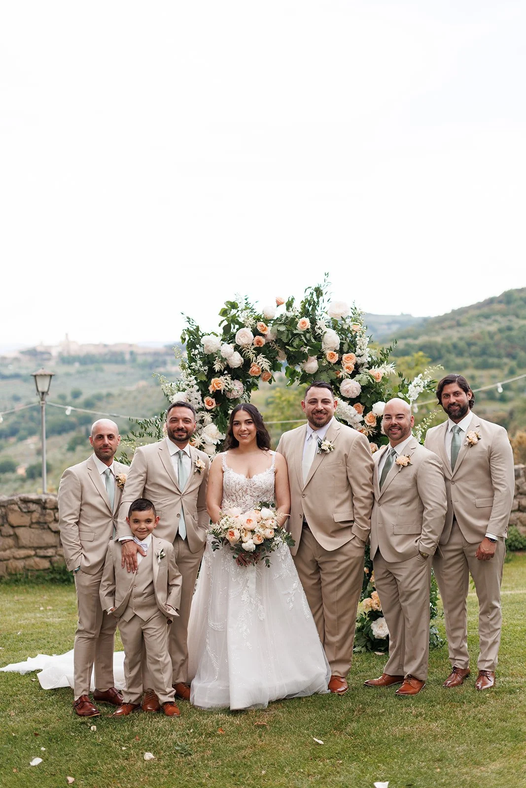 A wedding party standing outdoors in front of a floral arch with mountains in the background. The group includes a bride in a white lace gown holding a bouquet and six men in beige suits.