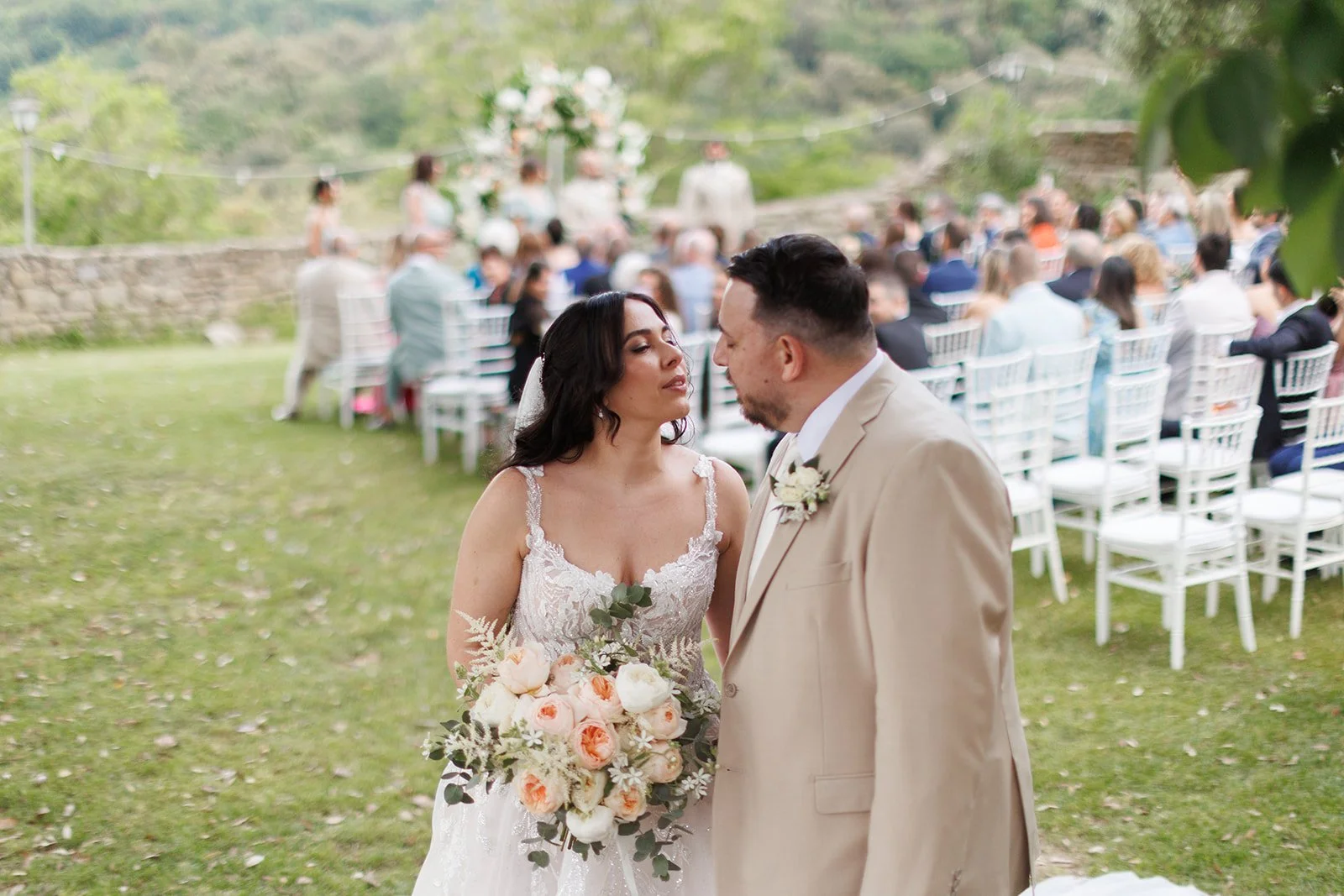 Bride and groom standing close together during their outdoor wedding ceremony, with guests seated behind them and a floral arch in the background.